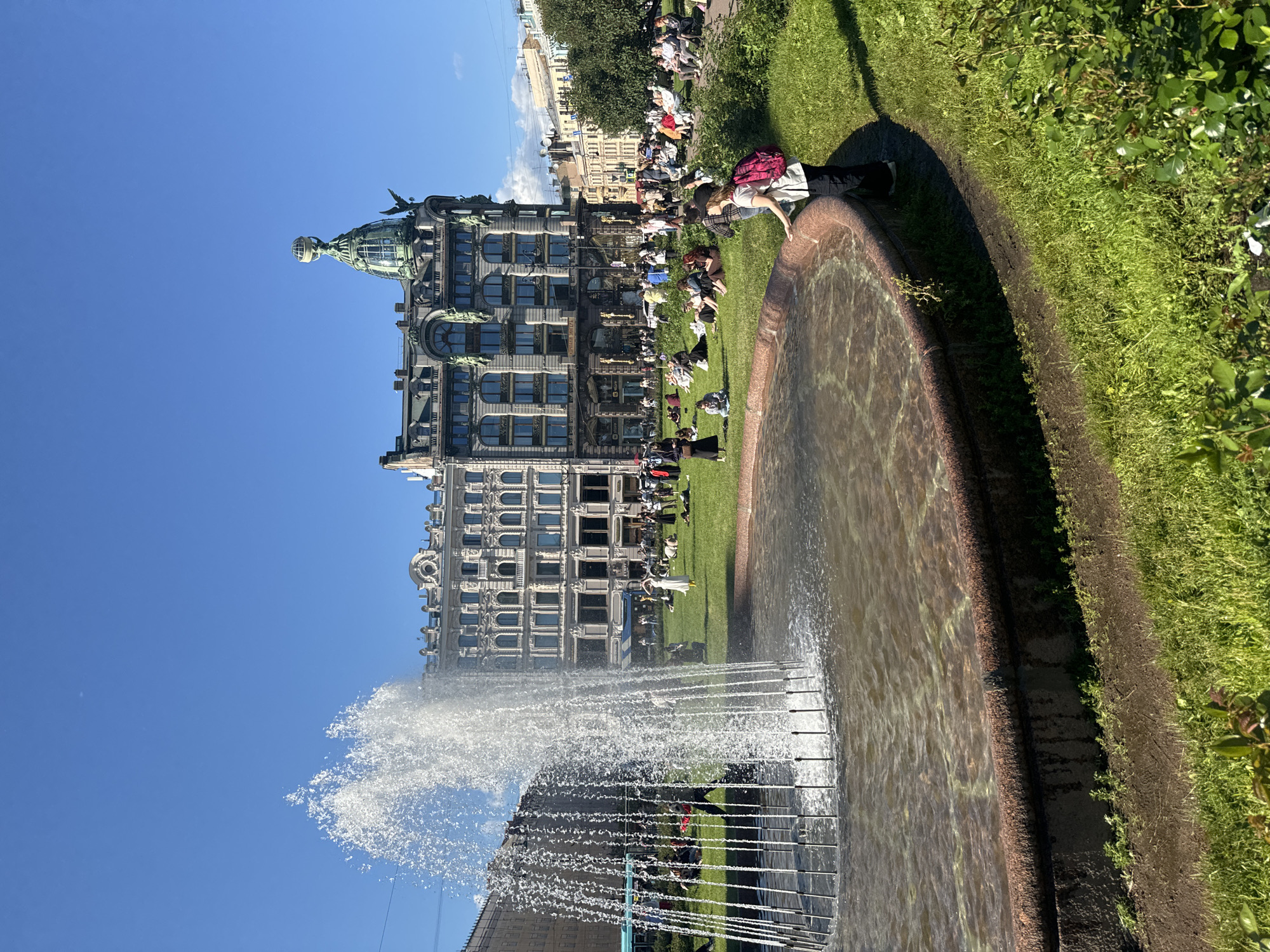 A wide shot of a public square in Saint Petersburg, Russia, featuring a large circular fountain in the foreground, its water spraying upwards. The fountain's edge is lined with a reddish-brown stone rim. Beyond the fountain, a lush green lawn is populated with numerous people, many of whom are sitting or lying down, enjoying the sunny day. In the background, a grand, ornate building with distinctive architectural details stands prominently. The sky is a clear, bright blue.