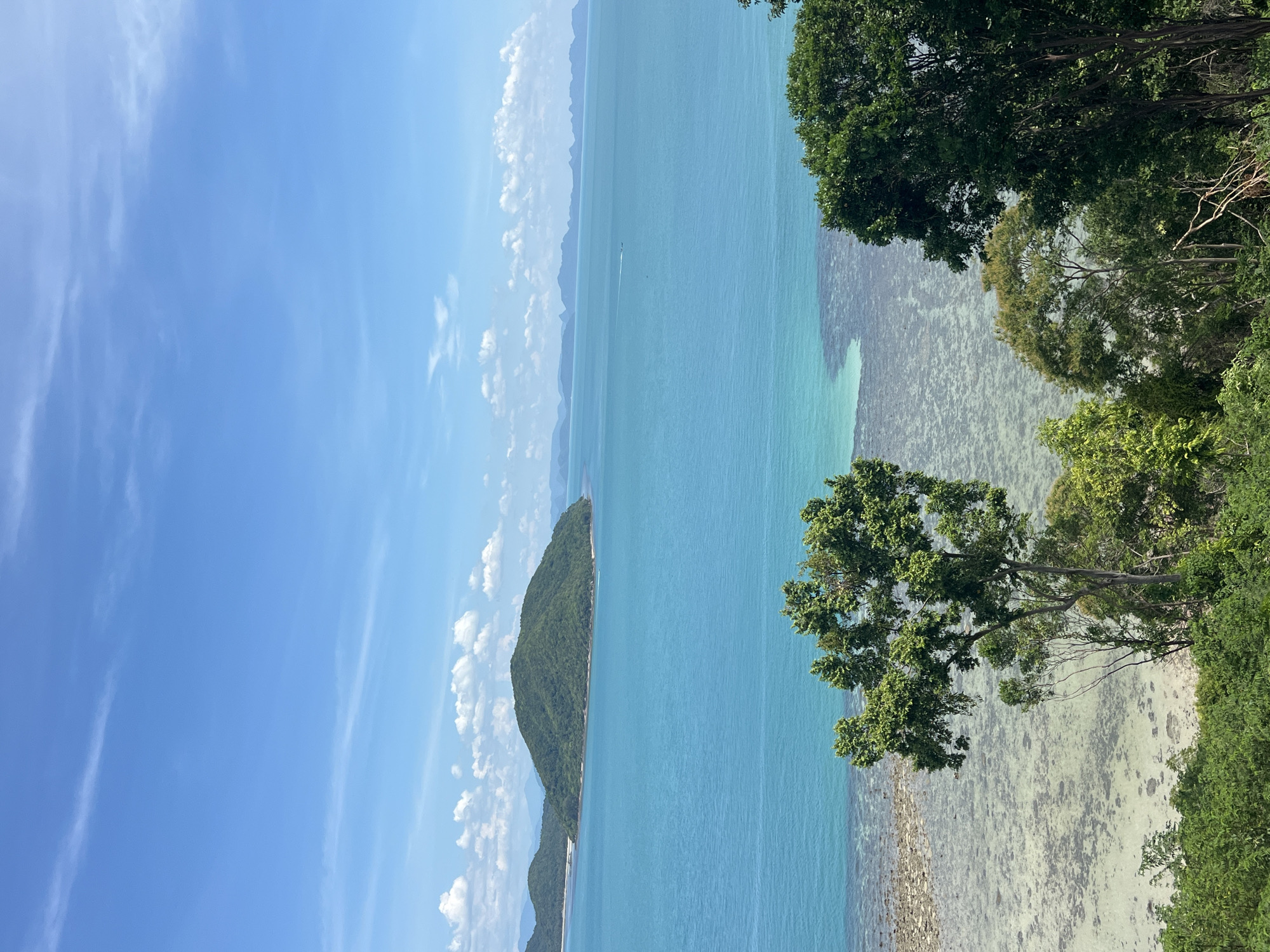 The image depicts a scenic view of the ocean from a higher vantage point, likely overlooking Baan Thong Krut, Thailand. The sky is a clear, bright blue with scattered white clouds. The ocean water is a vibrant turquoise, becoming lighter and more translucent closer to the shore, revealing the seabed with rocks and coral formations. In the middle distance, a lush green, rounded island rises from the water. Further in the background, a chain of hazy mountains can be seen on the horizon. In the foreground, the scene is framed by dense green foliage and trees. The overall impression is a tropical, serene coastal landscape under a sunny sky.
