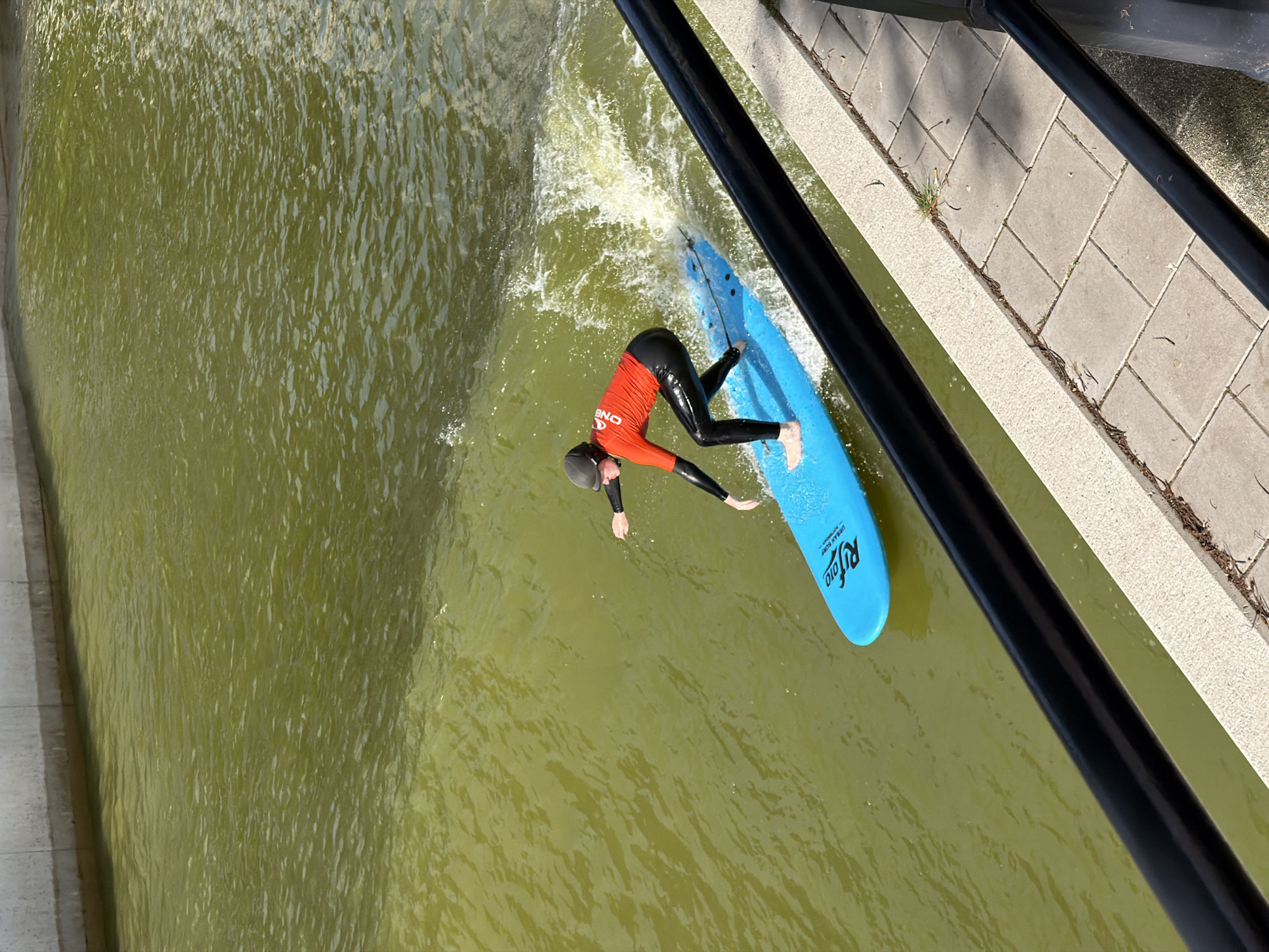 In Rotterdam, Netherlands, at the Zeilschip on the Maas, a person is shown surfing on a bright blue surfboard in a canal. The surfer, wearing a red top with white lettering that reads "O'NEILL," and black wetsuit pants, is actively engaged in surfing, with water splashing around the board. The canal water appears murky green, and a paved walkway with decorative tiles runs alongside it, bordered by black railings. The scene captures a moment of urban watersport activity.