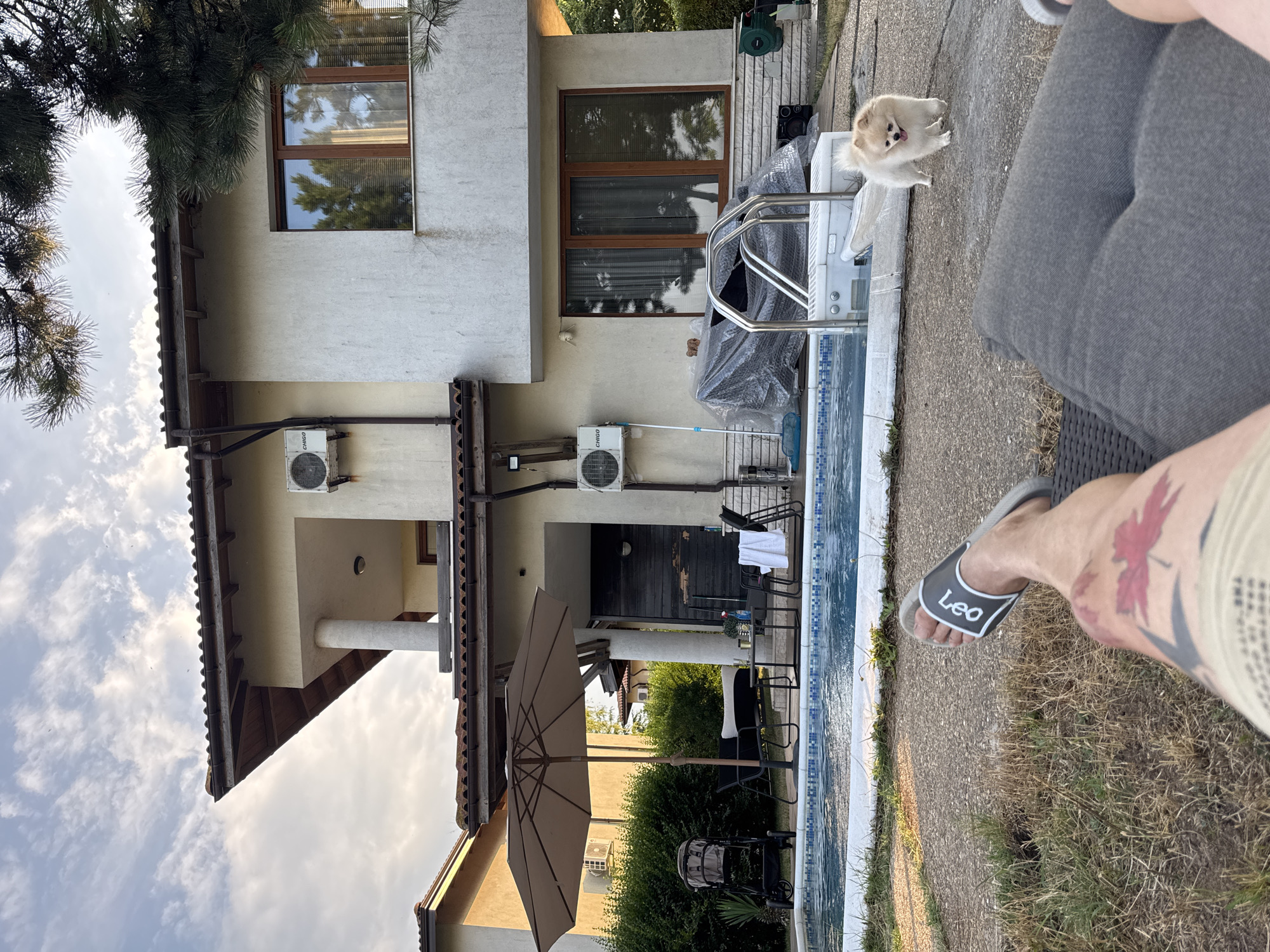 The image depicts a backyard scene in Balchik, Bulgaria, featuring a swimming pool and a multi-story house. A small, fluffy, light-colored dog stands on the concrete patio near the pool. In the foreground, a person's legs are visible, wearing grey slide sandals with the text "Leo" on them, and shorts with a tattoo of a red maple leaf on the thigh.
The house is light-colored with brown-trimmed windows and visible air conditioning units on its exterior. Adjacent to the house, a pool with blue tiled edges and a silver ladder is partially visible. A portion of a light brown umbrella is open, casting a shadow on the patio. Various outdoor furniture, including chairs and a small table, are arranged near the back of the house, some covered with plastic. A baby stroller is also present in the backyard. Green bushes and trees surround the property, suggesting a summer day. The sky is partly cloudy, with a mix of bright white clouds and patches of blue, indicating it is daytime, likely late afternoon or early evening given the light.