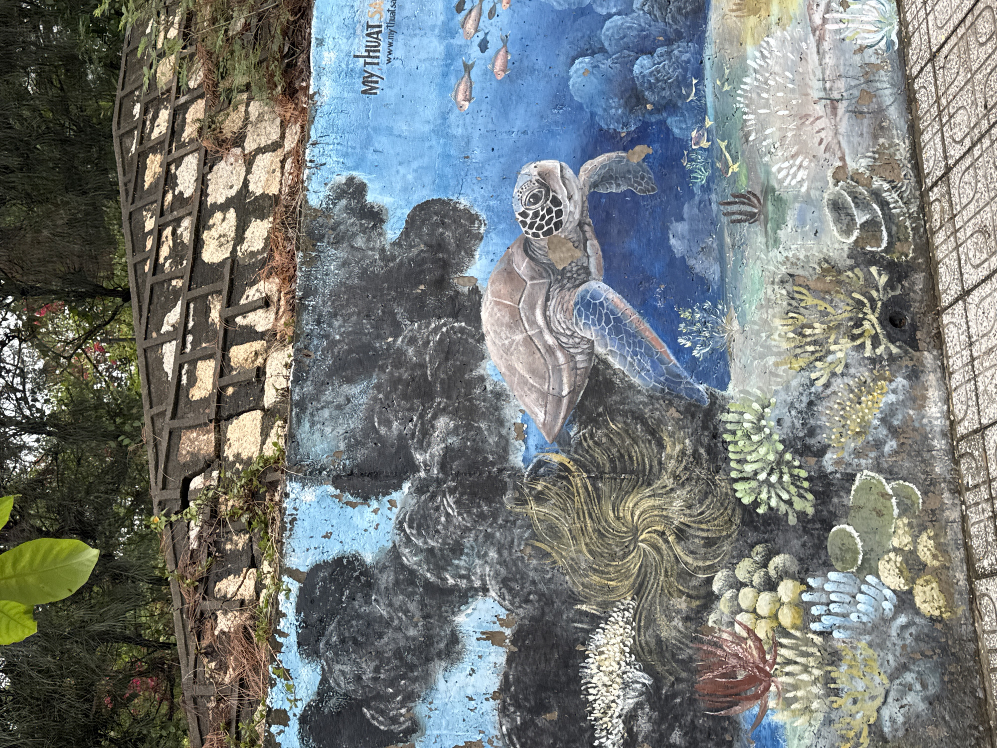 Here is a description of the image:
An eye-level, close-up shot of a marine-themed mural on an outdoor wall in the coastal city of Nha Trang, Vietnam. The artwork features a sea turtle prominently in the center, surrounded by coral and fish. The color palette is dominated by blues, browns, and greens, creating an underwater scene. A trellis wall structure is visible on the left side, where some greenery can be seen growing. The wall is rough and weathered, hinting at its outdoor location and exposure to the elements. Next to the right side of the mural, a pedestrian barrier is seen.
