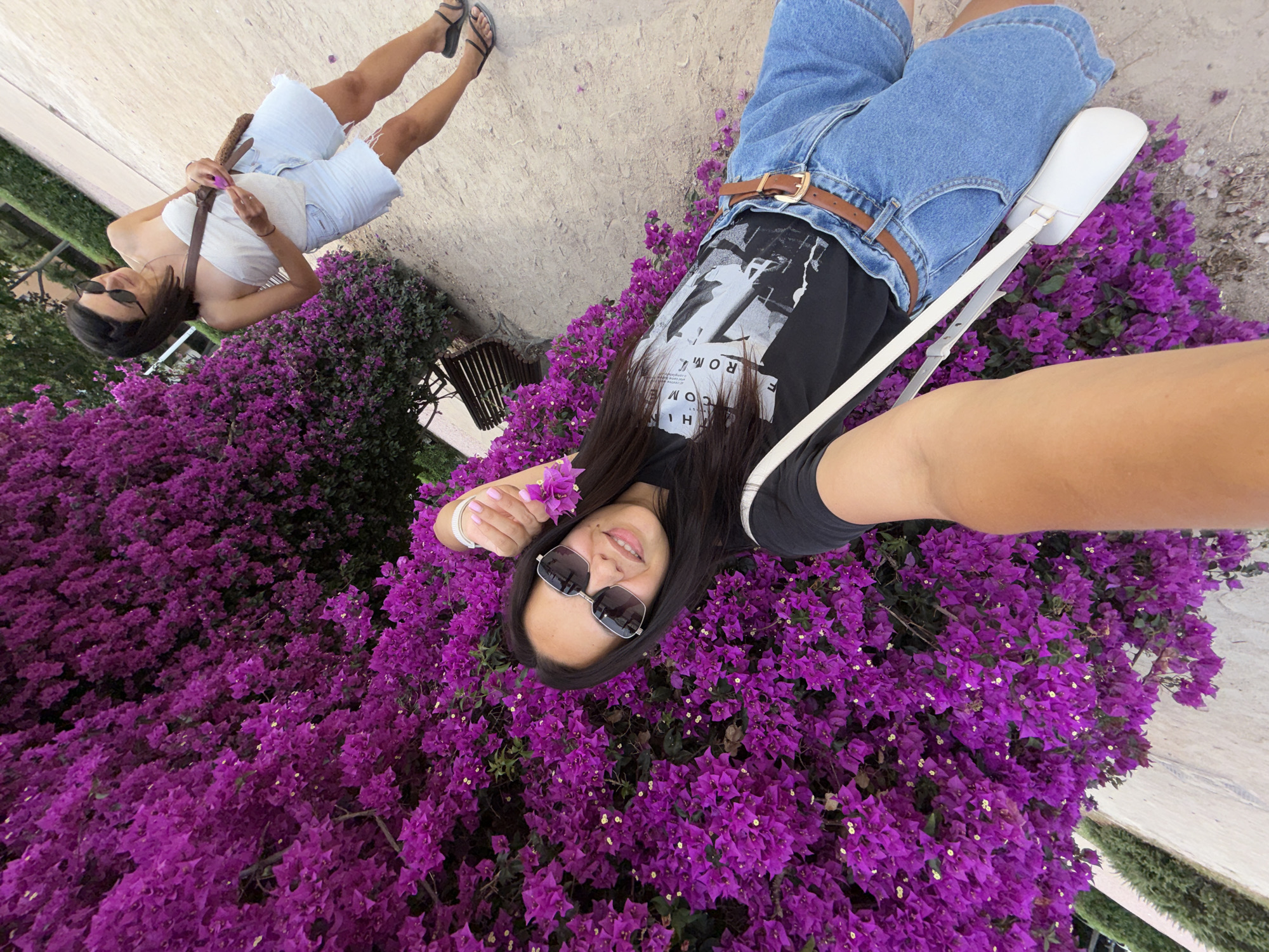 In an outdoor setting, likely a park or garden, two women are visible. The woman in the foreground is taking a selfie, holding a purple bougainvillea flower. She is wearing a black t-shirt with a graphic print, denim shorts, a brown belt, sunglasses, and a white crossbody bag. The woman in the background, slightly out of focus, is wearing a white strapless top, denim shorts, and black sandals. She is carrying a brown bag over her shoulder and appears to be looking to her left. The background features a large bush of vibrant purple bougainvillea flowers and a sandy path. A wooden bench is also visible behind the foreground woman. The lighting suggests it is daytime, possibly late afternoon.