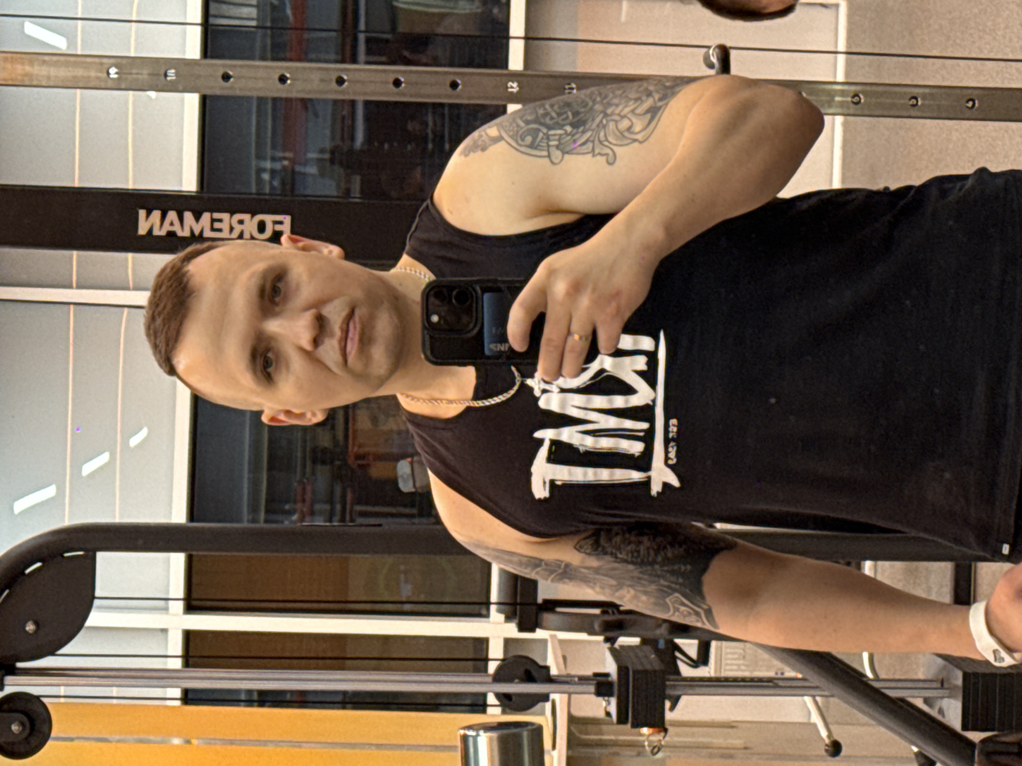 A man is taking a selfie in a gym, which appears to be located in Khimki, Russia, based on the backwards spelling of "Khimki" on the Foreman machine behind him. He is wearing a black tank top with a white graphic on it and a silver chain. He has tattoos on his left arm. He is holding a black phone in his right hand. The gym equipment behind him includes a weight machine with adjustable weights. The gym appears to be well-lit with natural light coming in from the windows.