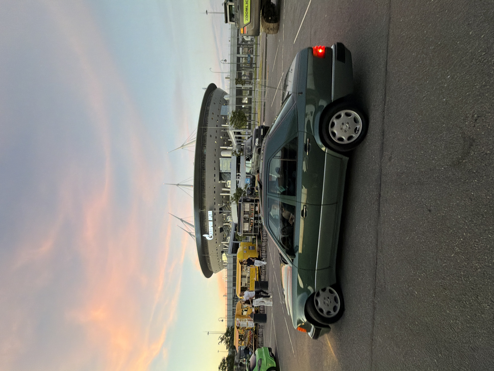 An eye-level, medium shot captures a dark green Mercedes-Benz sedan parked in a paved lot, with a large stadium and food trucks in the background. The car is angled slightly towards the viewer, with its rear brake light illuminated. The stadium, identified by Russian text on its facade, is a prominent architectural structure with a distinctive roof design. To the left of the stadium, several yellow food trucks are visible, along with a few people standing near them. The sky above is a soft gradient of pink and blue, indicating it is either sunrise or sunset. The ground is a textured asphalt surface, showing faint lines that suggest parking lot markings. In the distance, a construction vehicle, possibly an excavator, is also present. The scene appears to be an outdoor gathering or event, possibly a food festival or pre-game gathering at the stadium.