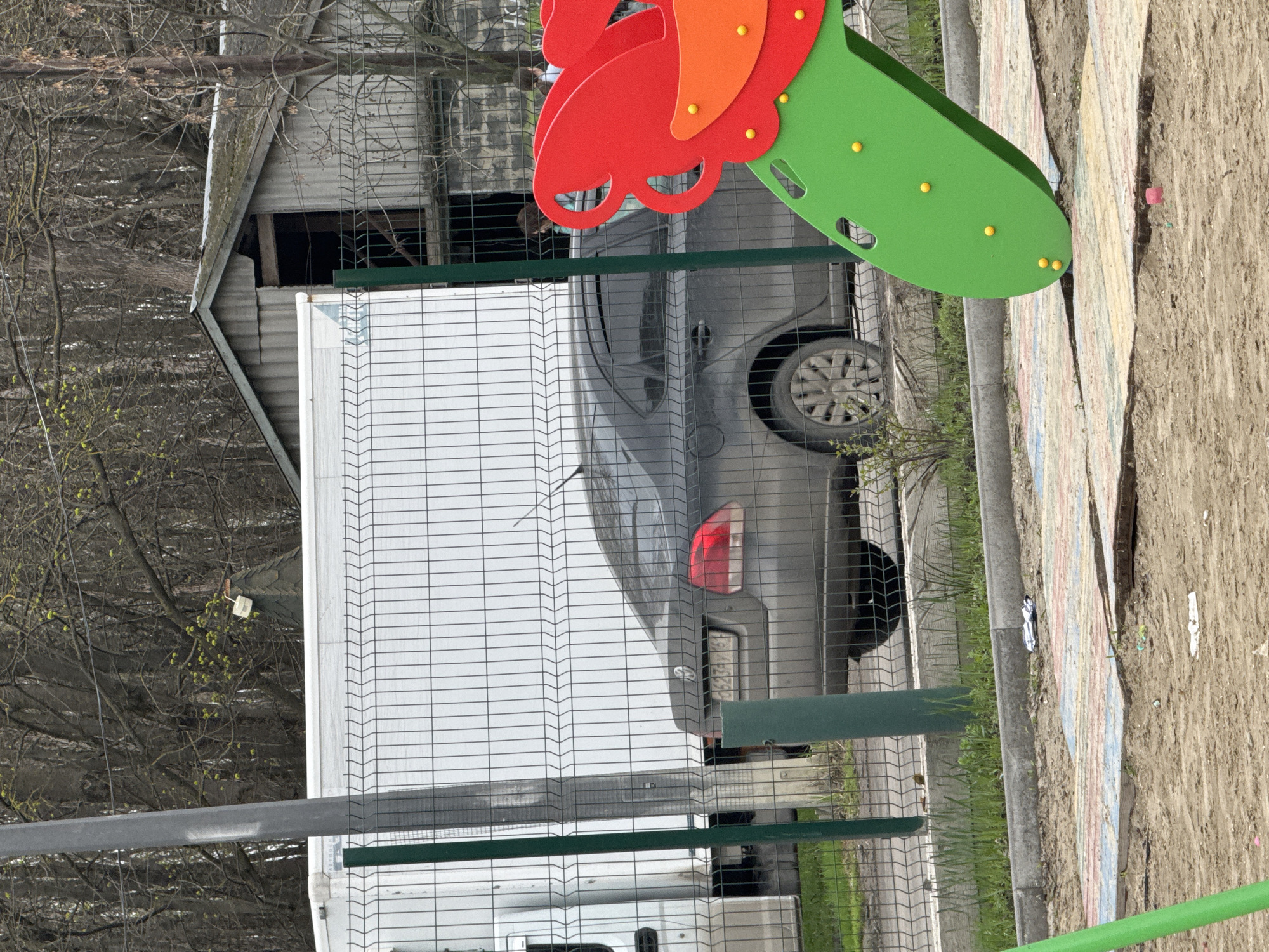 The image shows a fenced area with a car parked behind a white truck. In the background, there is a dilapidated shed or building. A colorful playground structure is visible in the foreground. The scene is likely in Rostov-on-Don, Russia. The focus is on the vehicle and the surroundings, giving a sense of the urban environment.