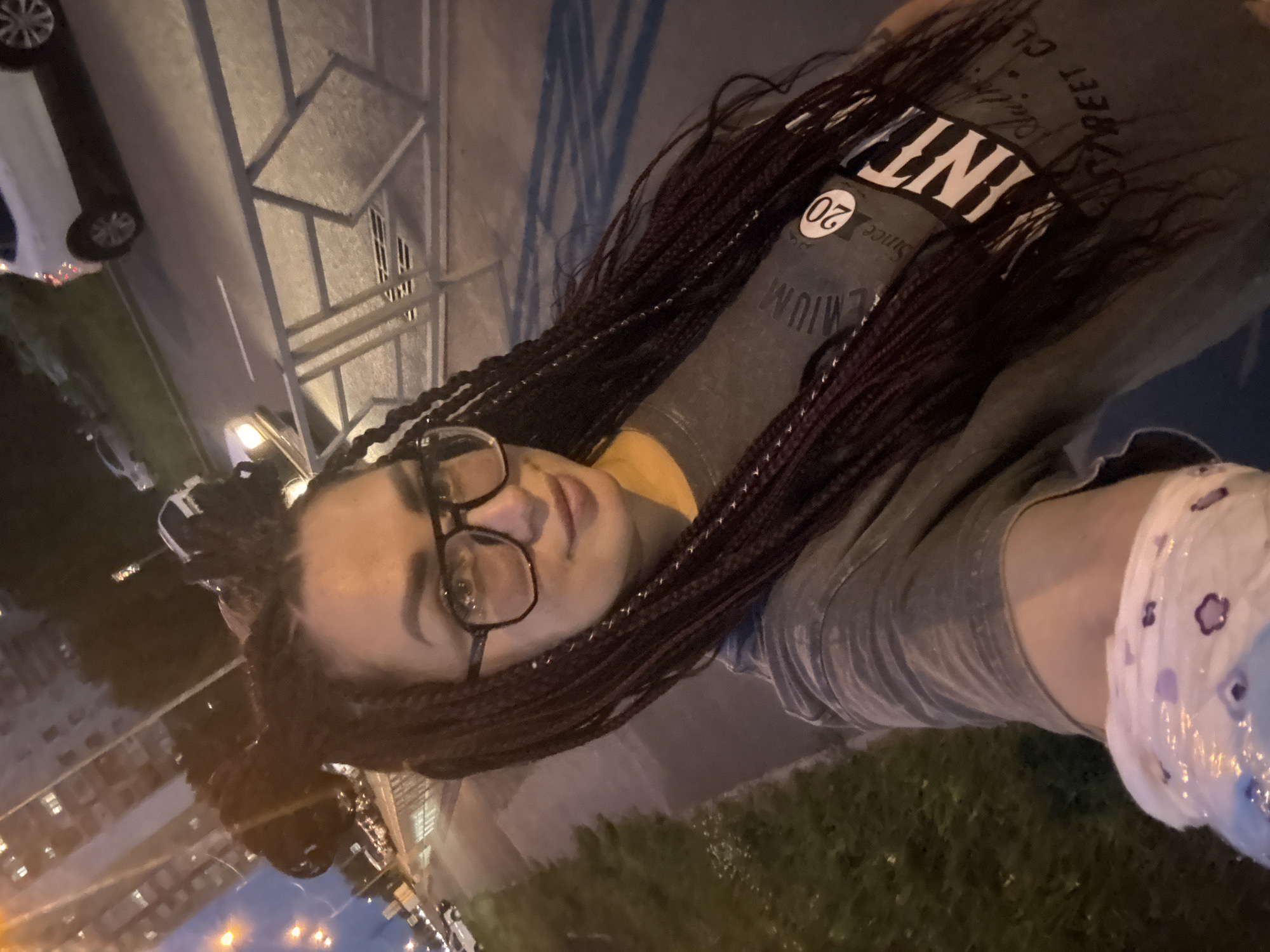 A woman with long, dark braided hair is taking a selfie on a sidewalk at night in Tolyatti, Russia. She is wearing glasses and a grey t-shirt with a graphic design. In the background, there are streetlights, cars, a building, and a metal fence. The sidewalk is next to a road.