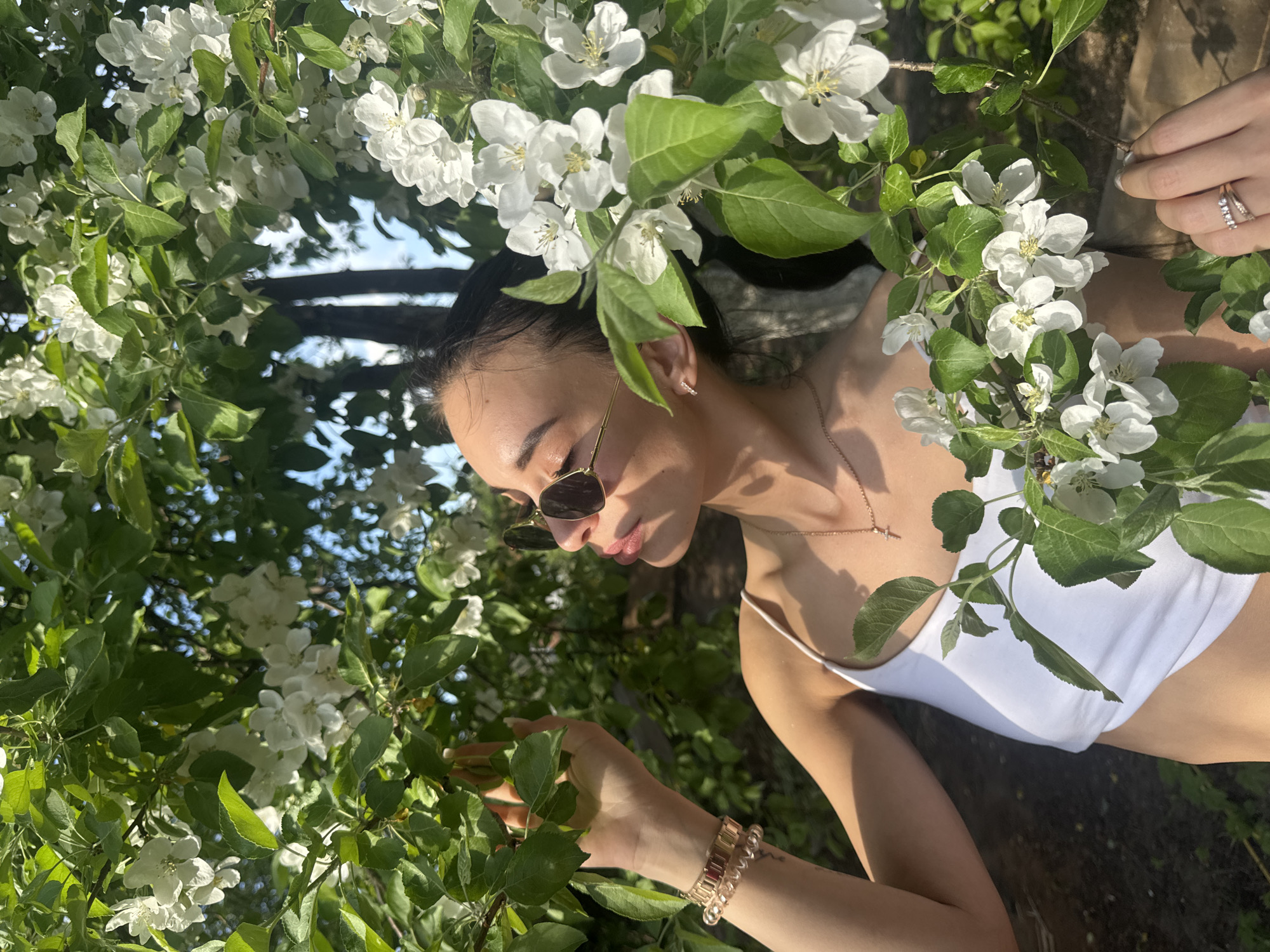 A young woman stands amidst blooming branches, likely apple blossoms, in the city of Chaikovsky, Russia. She wears a white tank top, sunglasses, and jewelry. The bright daylight illuminates the scene and casts shadows through the foliage. Her hands touch the leaves and flowers.