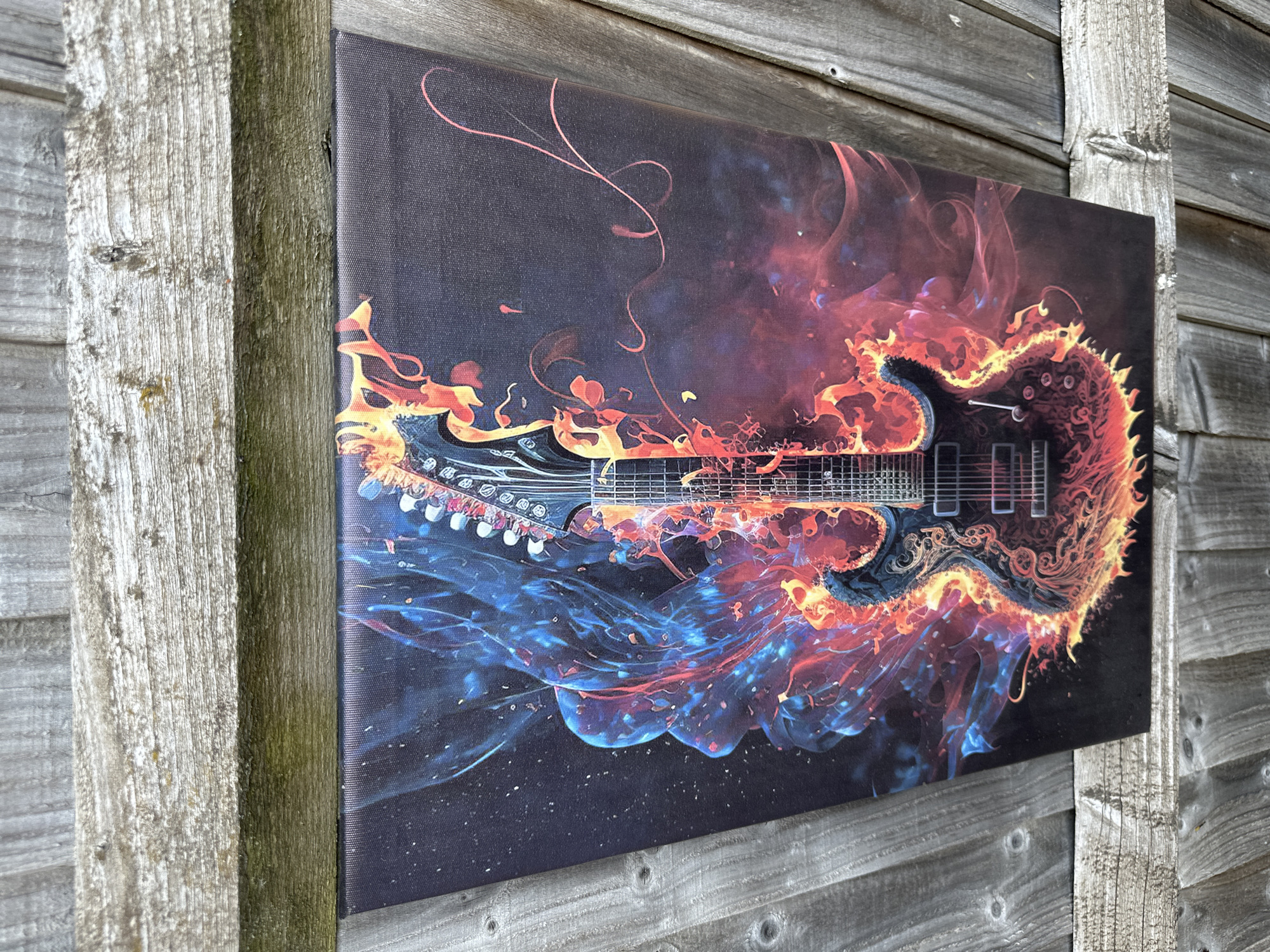 The image shows a canvas print of a stylized electric guitar engulfed in blue and orange flames, set against a dark background with specks of light resembling stars. The canvas is leaning against a weathered wooden fence. The fence consists of horizontal wooden planks with visible grain and knots. The canvas print itself is vibrant, with the guitar's details clearly defined amidst the fiery and smoky effect. The overall scene is static, with no people or actions depicted. The location is identified as a garden or outdoor space with a wooden fence, consistent with a residential area in Rugby, United Kingdom.