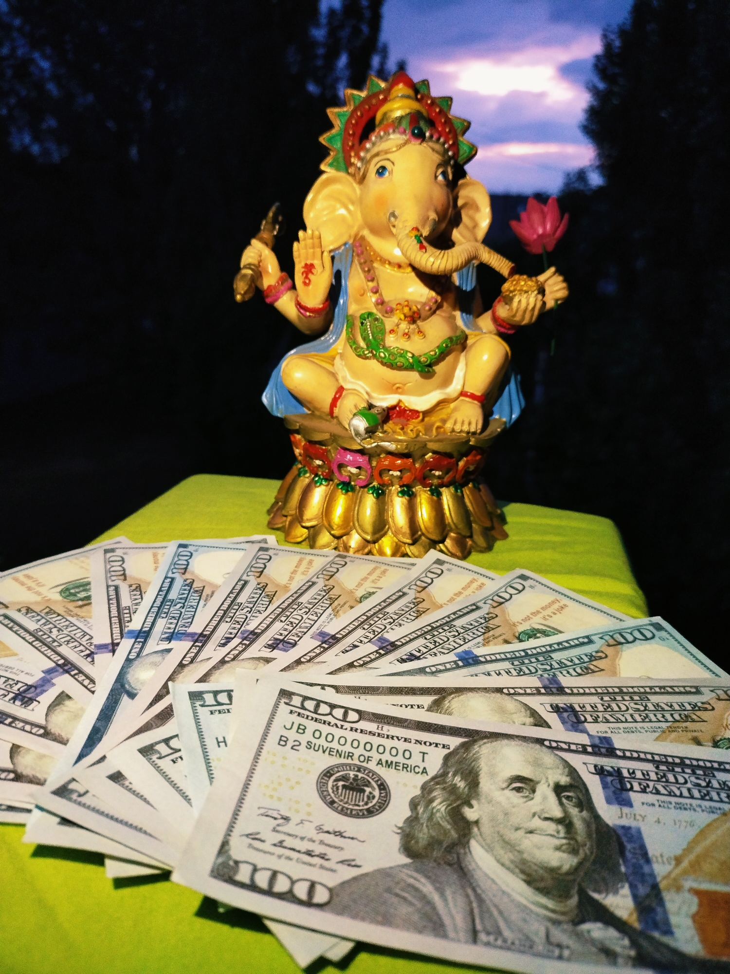 The image depicts a Ganesha statue and several US hundred-dollar bills arranged on a bright green surface. The statue is in the center, with its trunk, multiple arms, and ornate headdress clearly visible. It holds a lotus flower in one hand and a modak (sweet dumpling) in another. The hundred-dollar bills are fanned out in front of the statue, with Benjamin Franklin's portrait prominent on the visible bills. The background is dark, with hints of trees and a twilight sky with purple and orange hues. The scene is not located in Horishni Plavni, Ukraine, as there are no discernible Ukrainian landmarks or elements present in the image. The image appears to be a staged arrangement for symbolic or thematic purposes, possibly related to wealth, prosperity, or spirituality.