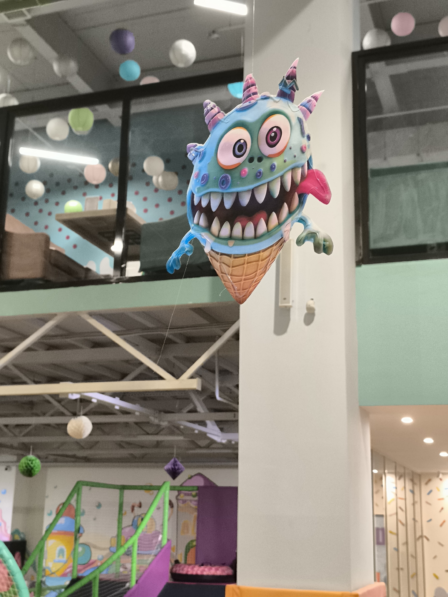 A whimsical, airborne decoration, resembling a cartoon monster with an ice cream cone for a body, hangs suspended from the ceiling in what appears to be an indoor play area. The monster is blue with pink and purple accents, featuring large, wide eyes, a gaping mouth filled with sharp teeth, and a bright pink tongue sticking out. Its arms are small and blue. The background reveals a colorful and playful environment, with suspended paper lanterns and decorative elements on the walls. The play area also includes climbing structures and soft play surfaces, suggesting it is a children's entertainment venue. The image does not provide any information that would allow for its location to be identified within the city administration of Qarağandı city, Kazakhstan.