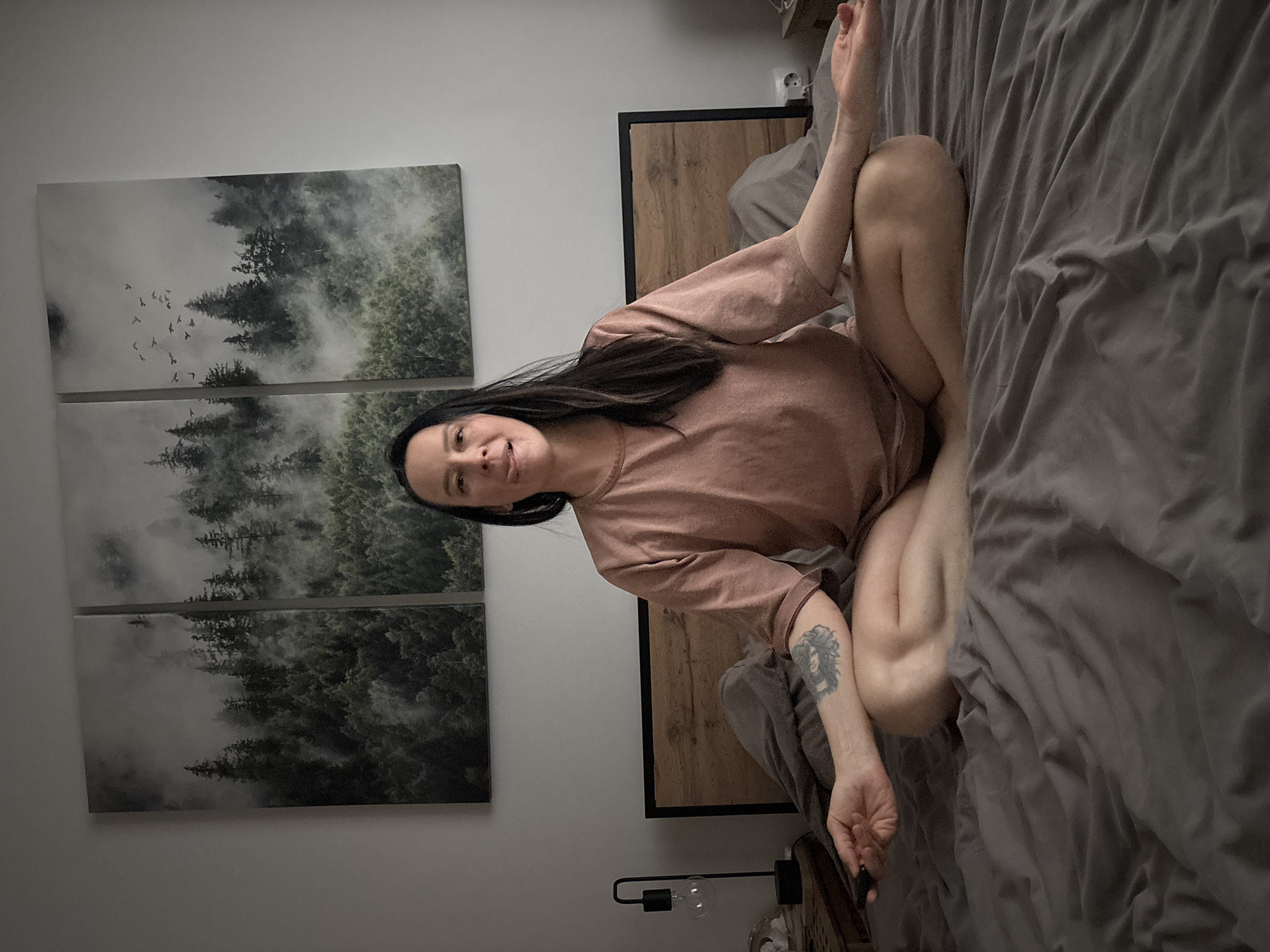 This media file captures an indoor scene, likely a bedroom, with the entire image rotated 90 degrees counter-clockwise.
The central subject is an adult woman with long dark hair, positioned in the middle of the frame, angled towards the left. She is wearing a loose-fitting, long-sleeved top in a light brown or dusty rose hue. Her legs are bent, and she sits in a relaxed, cross-legged posture on a bed. A notable detail is a tattoo on her right forearm, depicting a stylized head of a woman with flowing hair. She gazes directly at the viewer with a gentle, serene smile.
To her right (which would be the top of the image if upright), a bed with a wooden headboard frame is visible, covered with dark grey, rumpled bedding. An electrical outlet with a white plug can be seen on the wall behind the bed. To her left (the bottom of the image if upright), a bedside table holds a black industrial-style lamp with an exposed bulb.
On the wall behind and to the left of the woman (the left side of the frame), three rectangular framed art pieces are stacked vertically. These artworks feature a misty forest landscape with tall trees and a flock of birds in flight, rendered in muted tones of green, grey, and white. The wall itself is a plain, light color.
The activity appears to be a relaxed moment, possibly resting or contemplative, given her posture and calm expression. The lighting is soft and ambient, suggesting an indoor setting without harsh direct light. There is no visible text in the image.