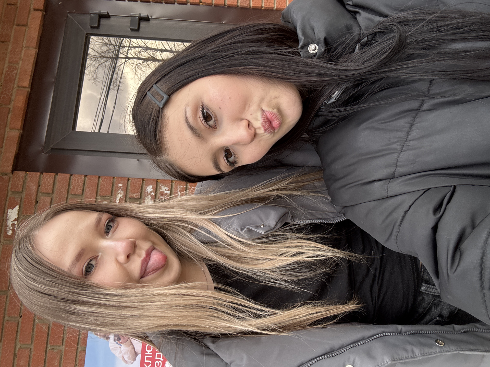 Two young women pose closely for a playful selfie, with the image rotated 90 degrees clockwise. The woman on the left has long, light blonde hair and light-colored eyes, smiling while playfully sticking her tongue out. She is dressed in a grey puffy jacket. Next to her, a partial poster or flyer is visible, displaying Russian text including "КЛЮ" and "3Д."
The woman on the right has long, dark hair, dark brown eyes, and makes a puckered, kissing face. She wears a dark grey or black puffy jacket and has a minimalist dark rectangular hair clip holding back part of her hair.
The background consists of a red brick wall on the left and a dark-framed window on the right. The window reflects bare tree branches silhouetted against an overcast, bright sky, indicating cool weather, possibly late autumn or winter. The overall mood is casual and cheerful. The context provided indicates the location is Фоки, Russia.