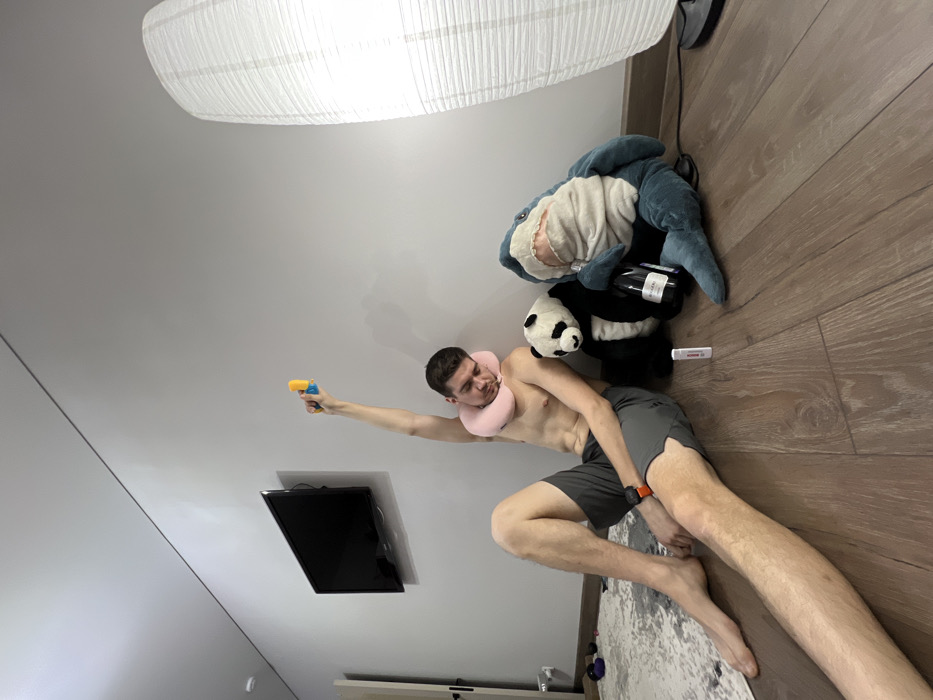 An indoor scene in Kazan, Russia, depicts a single adult male and two large stuffed animals on a wooden floor against a light grey wall. The male, shirtless and wearing grey shorts, has a light pink travel pillow around his neck and an orange-strapped smartwatch on his left wrist. He sits with his eyes closed, head tilted to his right, and his right arm extended vertically upwards, holding a small yellow and blue object. To his right, a black and white panda stuffed animal and a teal shark stuffed animal are positioned upright. The shark holds a dark-colored bottle with a white label between its fin and body. A white rectangular object with red markings, resembling a diagnostic test stick, lies on the floor in front of the shark. A black television is mounted on the wall behind the male. A tall, cylindrical white paper lantern floor lamp stands in the far right of the frame. A patterned light-colored rug is partially visible on the left side of the wooden floor.