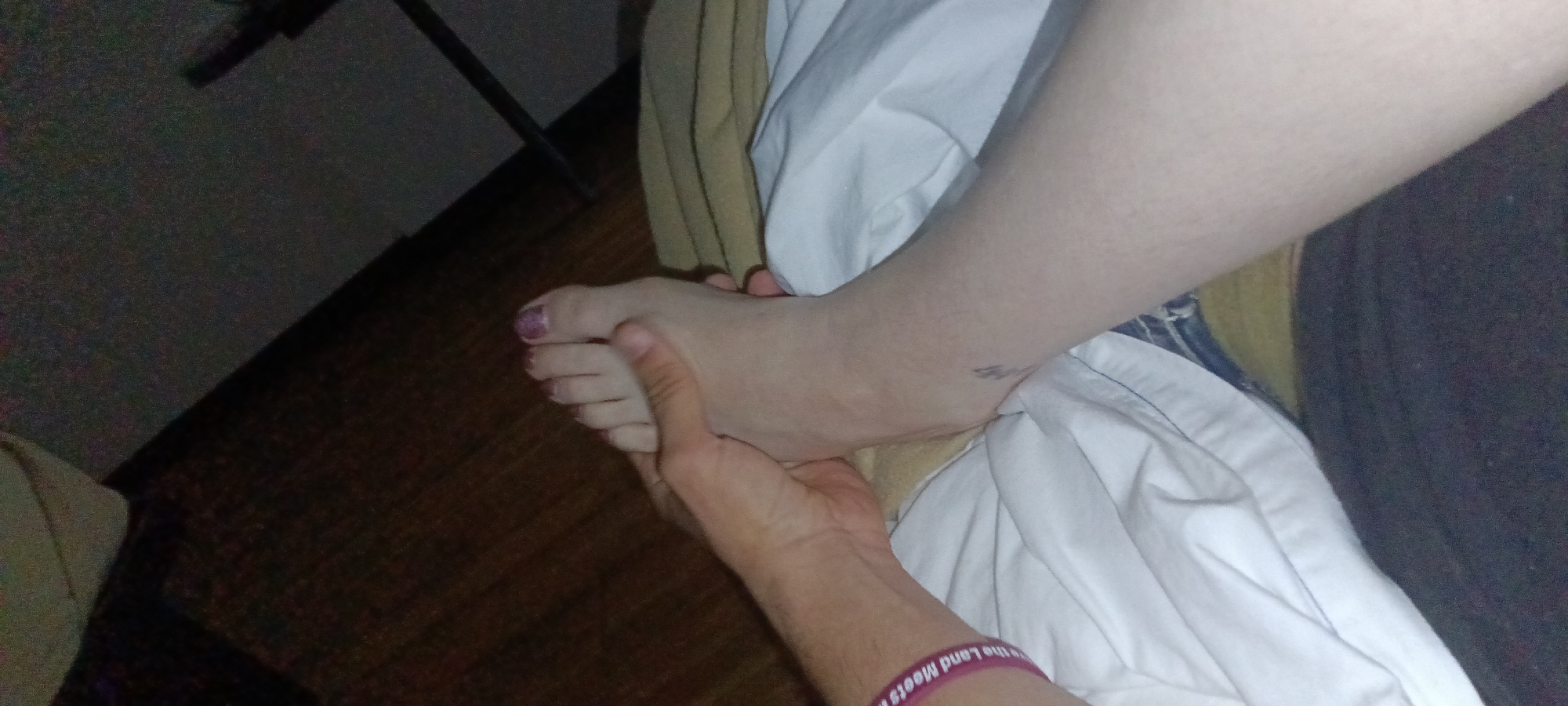 A foot is being held by a hand. The foot has purple nail polish on the big toe. The hand and foot are on white sheets and a tan blanket. A purple wristband is on the wrist. A wooden floor is visible. A black metal stand is visible in the background. It is dark in the room. It is not possible to determine the location in Fort Smith, United States of America from the image.