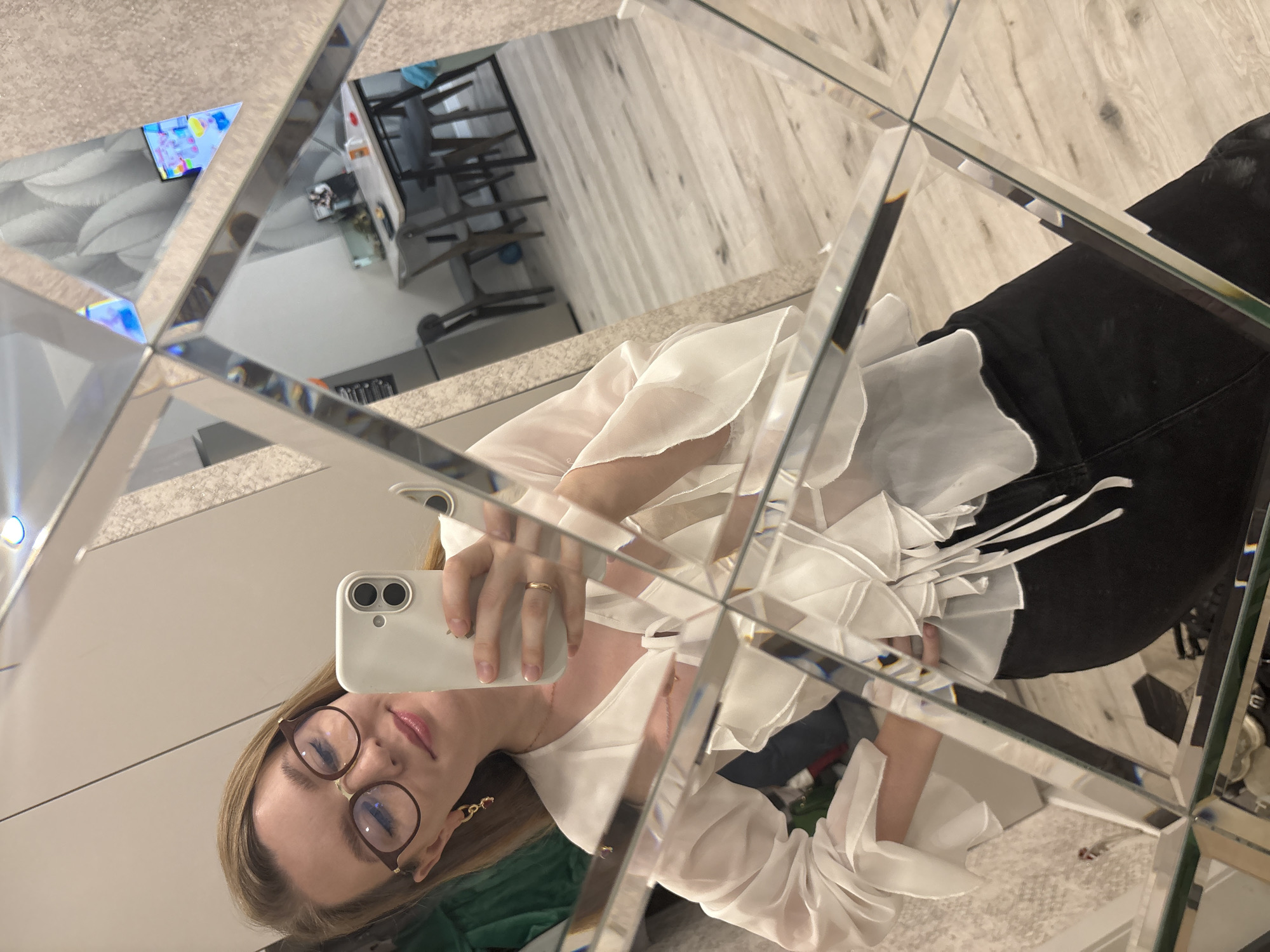 A young woman is taking a selfie in front of a multifaceted mirror. The mirror reflects the room behind her, showing a kitchen area with chairs and a glimpse of a television screen. The woman is wearing glasses, a white blouse with ruffles, and black pants. The location is Izhevsk, Russia.