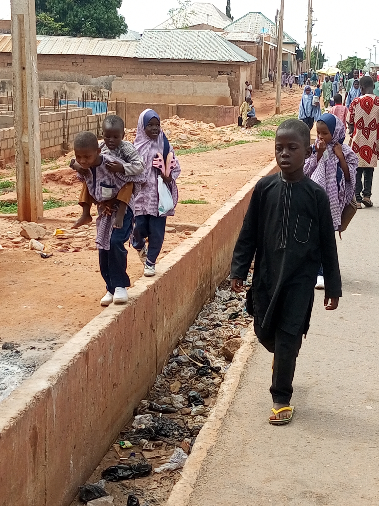 In Gombe, Nigeria, a group of children walks along a dirt road next to a concrete drainage ditch filled with debris. In the foreground, a young boy in a black traditional tunic and trousers walks towards the viewer, wearing yellow flip-flops. To his left, two younger children are walking. One is carrying the other on his back. The boy on the back is wearing a striped shirt and pants. The carrier is wearing a striped shirt and dark blue trousers. A girl with a hijab is walking behind them, holding a white plastic bag. In the background, other children and adults are walking down the road. The buildings on the left appear to be under construction, with exposed brickwork and scaffolding. The overall scene depicts a typical street in Gombe, showing daily life and the presence of children in a somewhat cluttered urban environment.