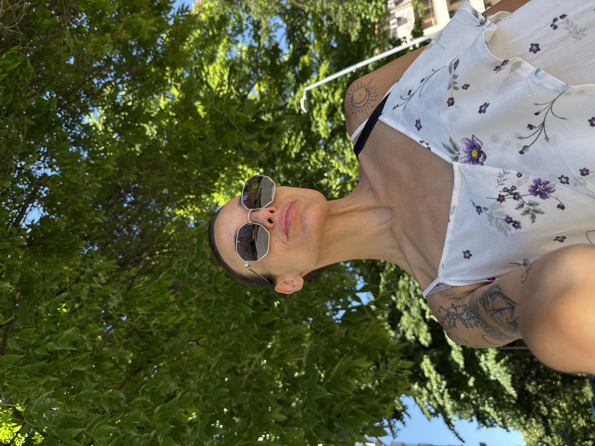 A woman is photographed outdoors, from the chest up, against a backdrop of lush green foliage and a bright blue sky. She is wearing sunglasses and a white dress adorned with purple floral patterns. Tattoos are visible on her right arm and shoulder. The image appears to have been taken in Pleven, Bulgaria, though no specific landmarks are identifiable.