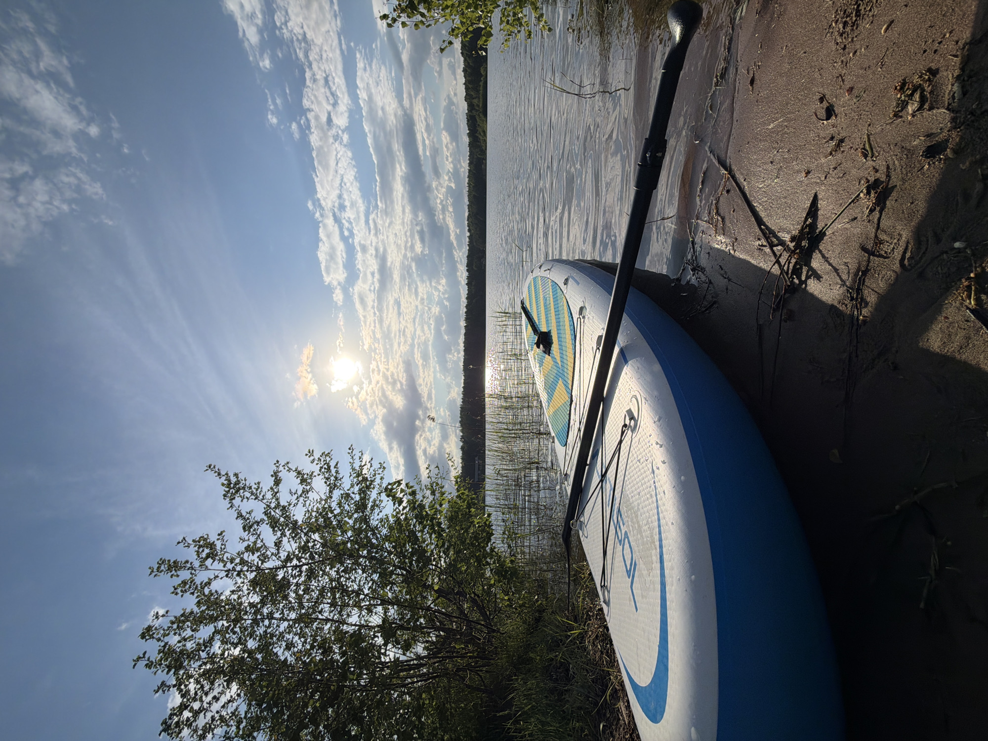 The image shows a paddleboard lying on a sandy shoreline next to a lake or river. The paddle is resting on the board. The sky is visible, with clouds and sunshine. Trees and vegetation are visible along the shoreline. The text on the paddleboard reads "105 SOL". It's impossible to determine the location is Selychka, Russia from the image alone.
