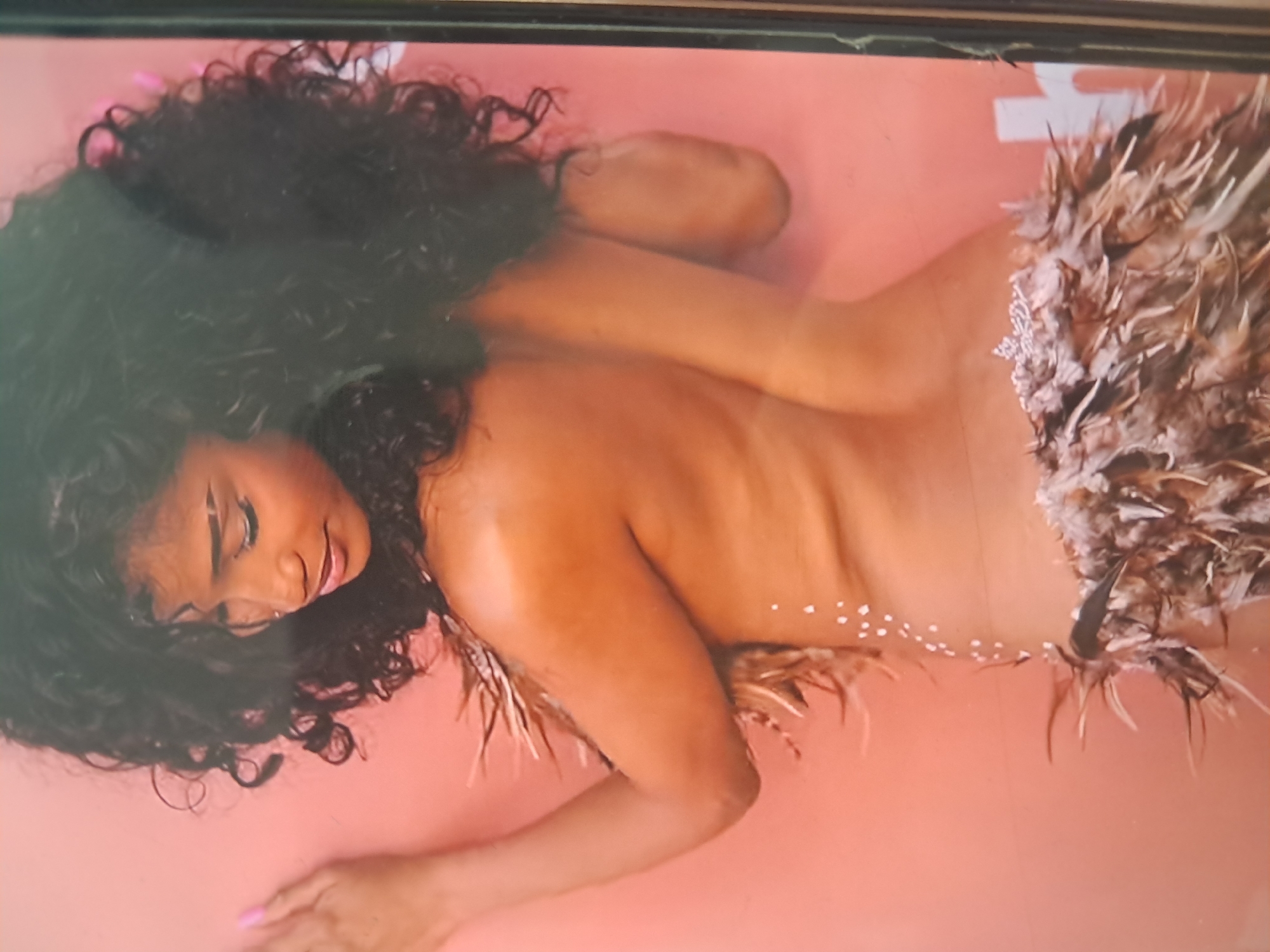 The media file shows a dark-skinned woman with long, curly dark hair lying on her stomach, facing away from the viewer, on a peach or light pink surface. She is wearing what appears to be a feathered or fluffy garment, possibly a skirt, that is light brown or beige in color and extends from her lower back/hips downwards. Small white embellishments, possibly beads or sequins, are visible on her lower back and on the garment near her hips. Her upper body is bare, revealing her back and shoulders. The woman's face is partially visible, tilted towards the viewer, and she has makeup on, including dark eyeliner or eyeshadow and pink lipstick. Her eyes are closed or looking downwards. One of her hands, with painted nails, is visible in the lower left of the frame. The overall impression is that of a posed photograph, possibly for an artistic or fashion context. The setting appears to be a studio with a plain colored background. There is no visible text. The time of day cannot be determined, and there are no specific location cues other than the implied indoor studio setting.