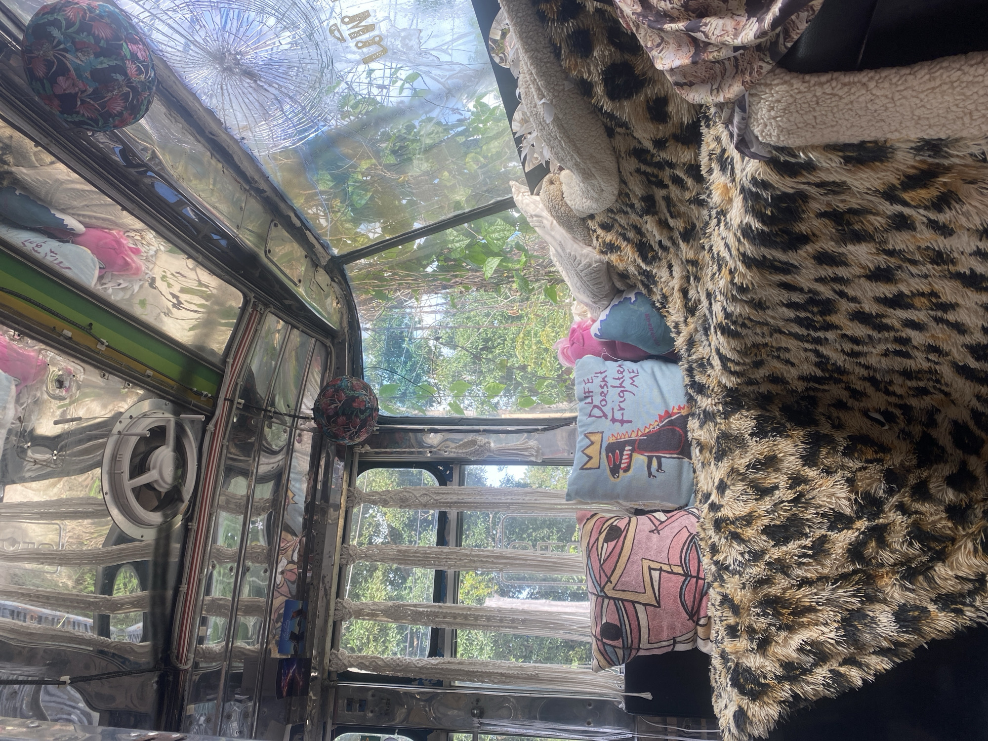 The image shows the interior of a vehicle, possibly a bus or jeepney, in Thong Sala, Thailand. The interior is adorned with patterned pillows, a leopard-print blanket, and reflective metal trim. The windshield is cracked. Outside, there's green foliage and trees.