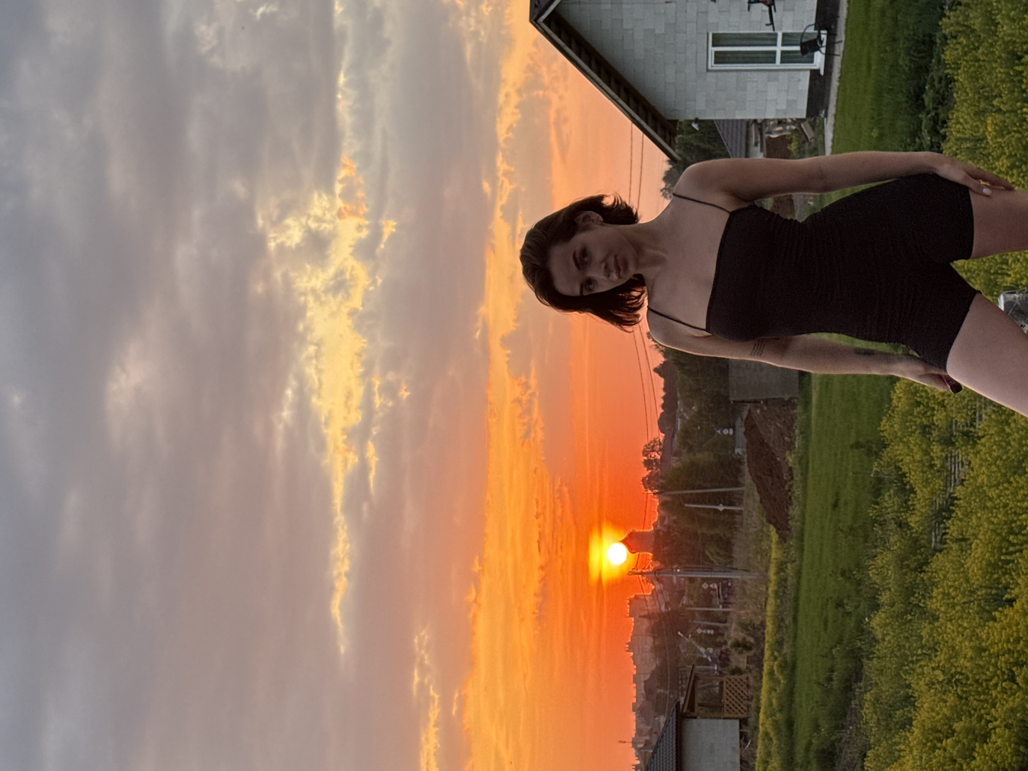 A woman stands in the outdoor setting of Лобаново, Russia. She has dark hair, and fair skin, and is wearing a black outfit with spaghetti straps. Behind her is an orange sunset on the left, and a house on the right. There is green foliage and vegetation in the lower right corner of the image.