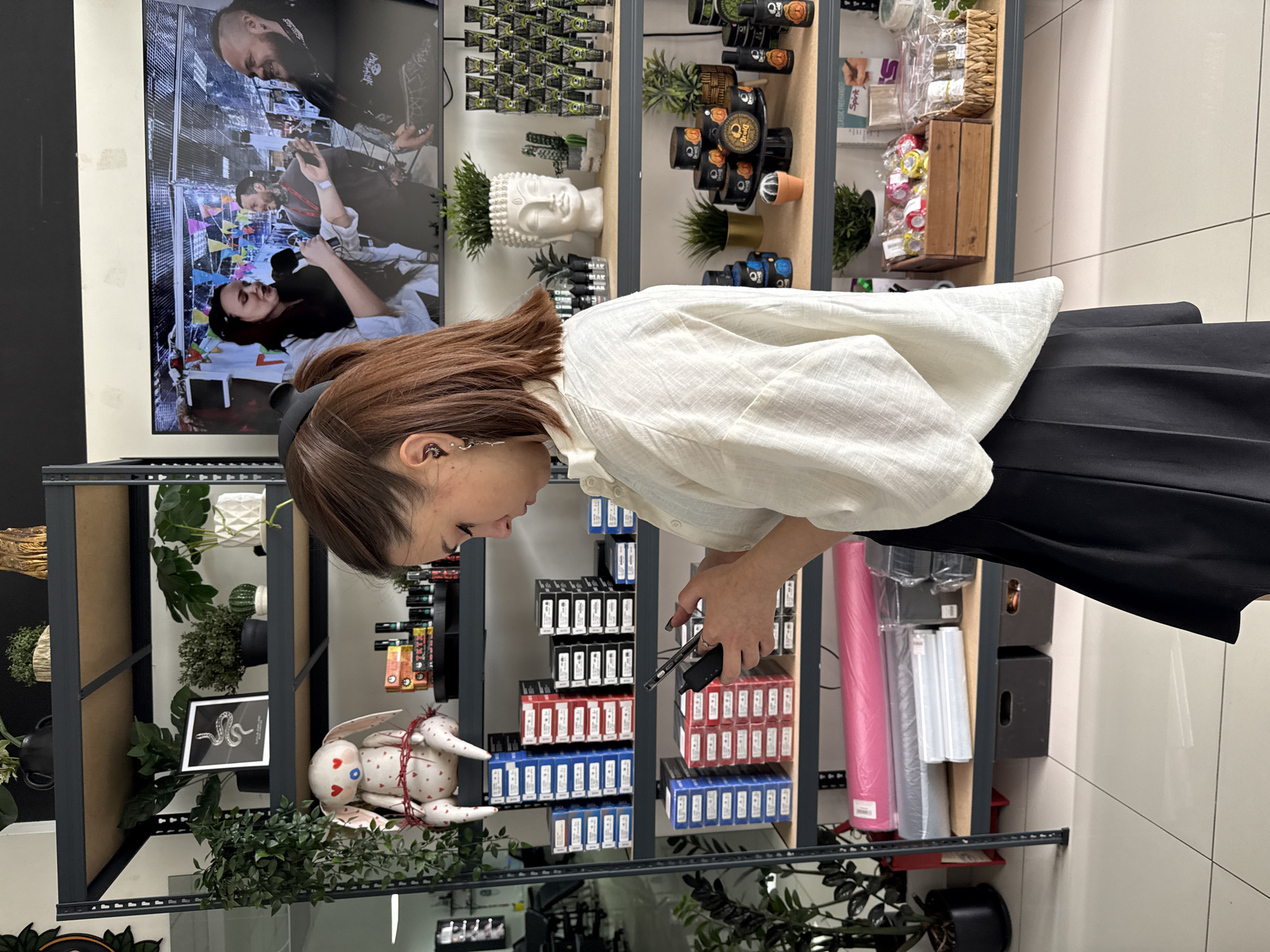 A young woman with brown hair is standing in front of a shelf full of various items, taking a photo with her smartphone. She is wearing a white blouse and a black skirt. The shelf contains items such as plants, a Buddha head, candles, and packaged goods. There's also a TV screen showing a recording or live stream in the background. On the lower shelves are boxed items and a stuffed rabbit toy. The location is a retail setting, potentially in Rostov-on-Don, Russia, based on the implied geographical constraints of the query. The woman is focused on capturing the image with her phone.