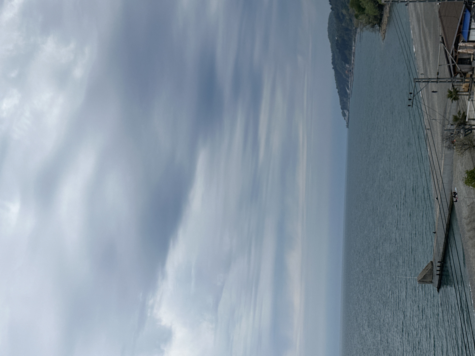 The image displays a coastal landscape captured on an overcast day. A large expanse of calm, blue-grey water, likely the sea, fills the central and lower portions of the frame, its surface showing subtle ripples. To the left, a distant coastline is visible, featuring lush, green hills or mountains that gradually recede into the hazy horizon.
On the right side of the water, a paved area or road runs parallel to the shore. Along this path, a series of utility poles are prominently featured, supporting numerous overhead power lines that crisscross the mid-ground. A small, triangular concrete structure, possibly a pier or breakwater, extends into the water from the right edge, with a thin pole or mast visible at its tip. Further to the right, behind the power lines, some buildings or structures are partially visible, and a few palm trees are scattered in the foreground-right.
The upper part of the image is dominated by a cloudy sky, characterized by soft, elongated streaks of light grey and white against a lighter blue background, indicating an overcast daytime. No people are visible, nor is any discernible text present in the image. The overall scene conveys a peaceful and subdued atmosphere typical of a coastal area under cloudy conditions.