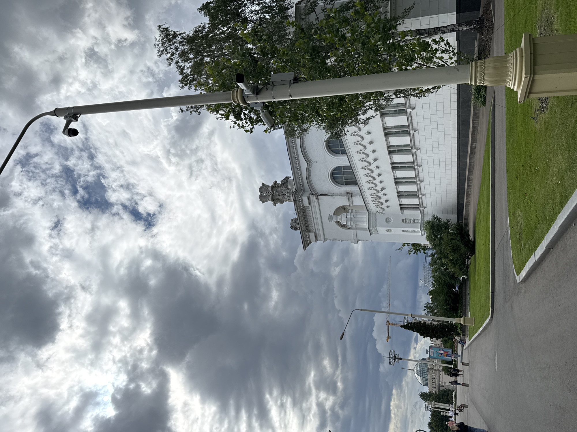 The image depicts a street scene in Moscow, Russia, with a focus on architectural elements and the surrounding environment. A tall, light-colored pole with attached security cameras and a cable runs diagonally across the frame. Behind it, a grand white building with arched windows and decorative detailing is visible. Trees with green foliage are interspersed with the architecture, adding a natural element to the urban setting. A paved walkway runs alongside a manicured lawn, suggesting a public park or exhibition area.
In the distance, several structures hint at ongoing development or ongoing events, including construction cranes and a dome-shaped building. The sky is filled with dramatic, heavy clouds, indicating overcast weather. The lighting suggests it is daytime, but the cloud cover creates a subdued atmosphere. People are present in the background, walking along the path and engaged in various activities, though they are small in the frame and not the primary focus. The overall impression is of a large, open public space under a moody sky. No discernible text is visible in the image.