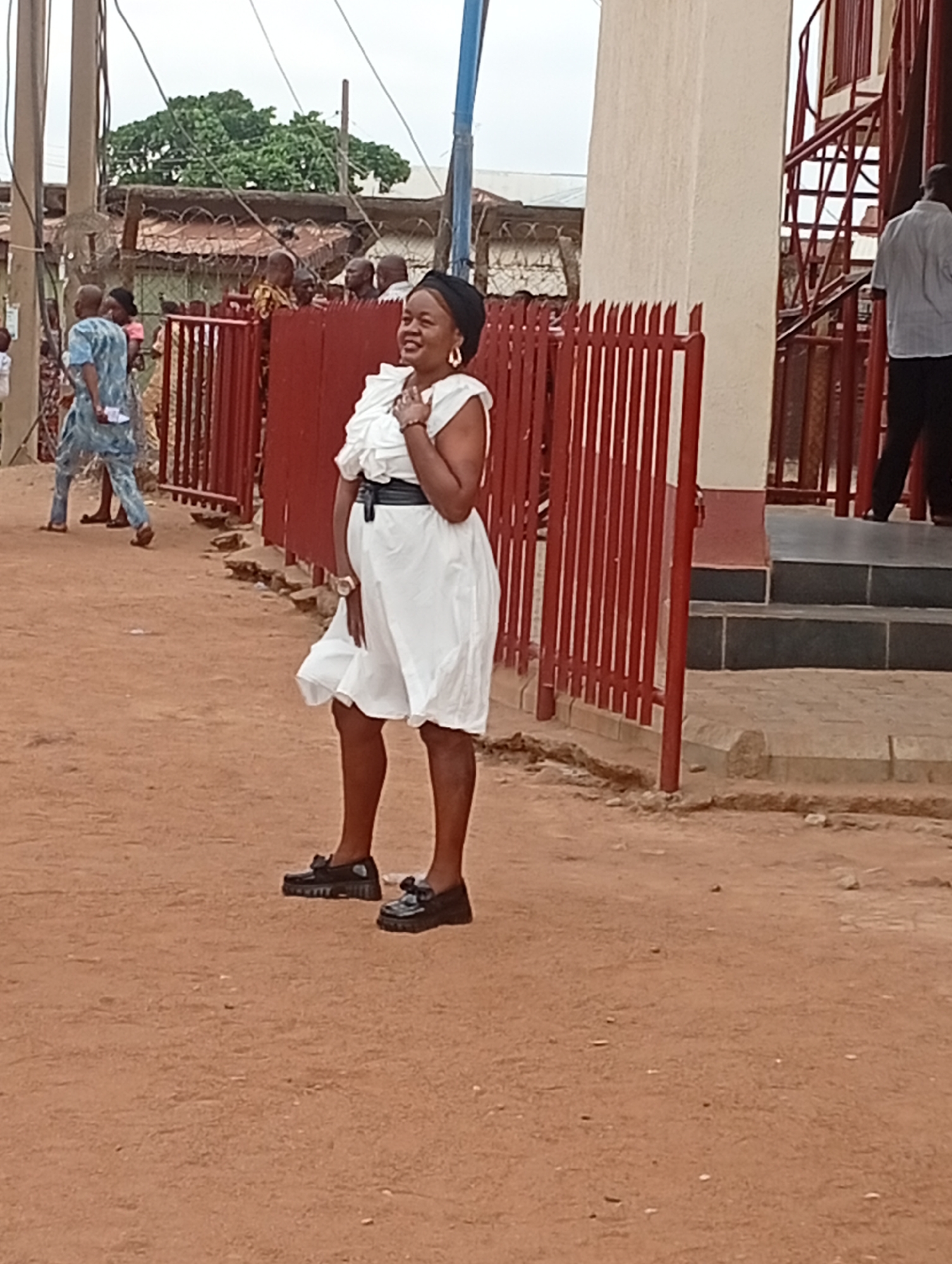The image shows a woman in a white dress, black shoes, and a black headwrap standing outside. She is smiling and has her hand on her chest. Behind her is a red metal fence and a building with a white column and a metal staircase. In the background, there are other people and a building with a corrugated metal roof. The ground is reddish-brown dirt. The location is identified as Narayi, Nigeria.