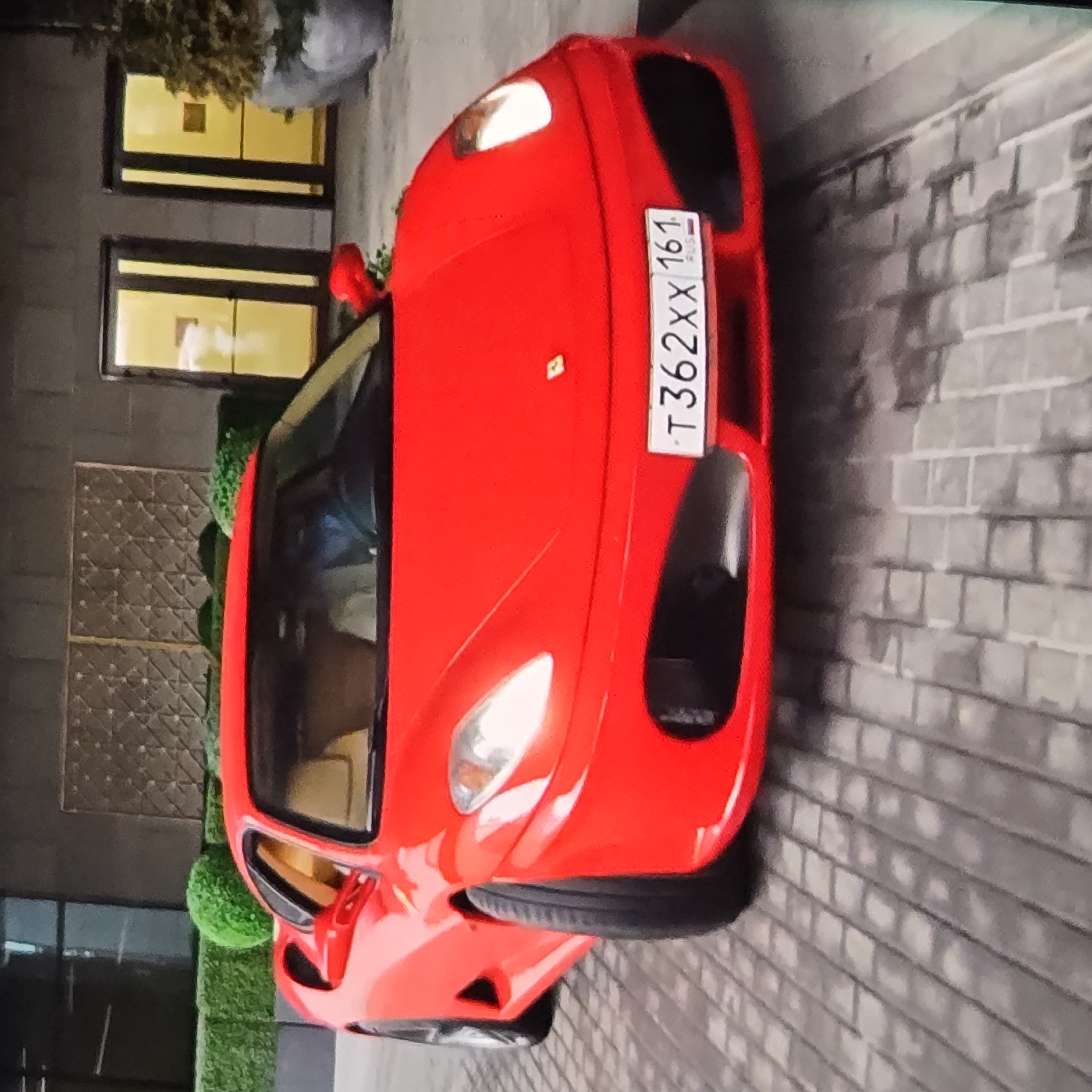 A red Ferrari F430 sports car is parked on a cobblestone driveway in front of a modern building with large glass doors and decorative metal screens. The car's license plate reads "T 362XX 161 RUS". The car has tan leather interior and is positioned at an angle, facing slightly to the right. There are neatly trimmed green hedges to the left of the car and a large potted plant on the right. The lighting suggests it is daytime, with sunlight casting shadows on the driveway. The building appears to be a commercial or residential complex.