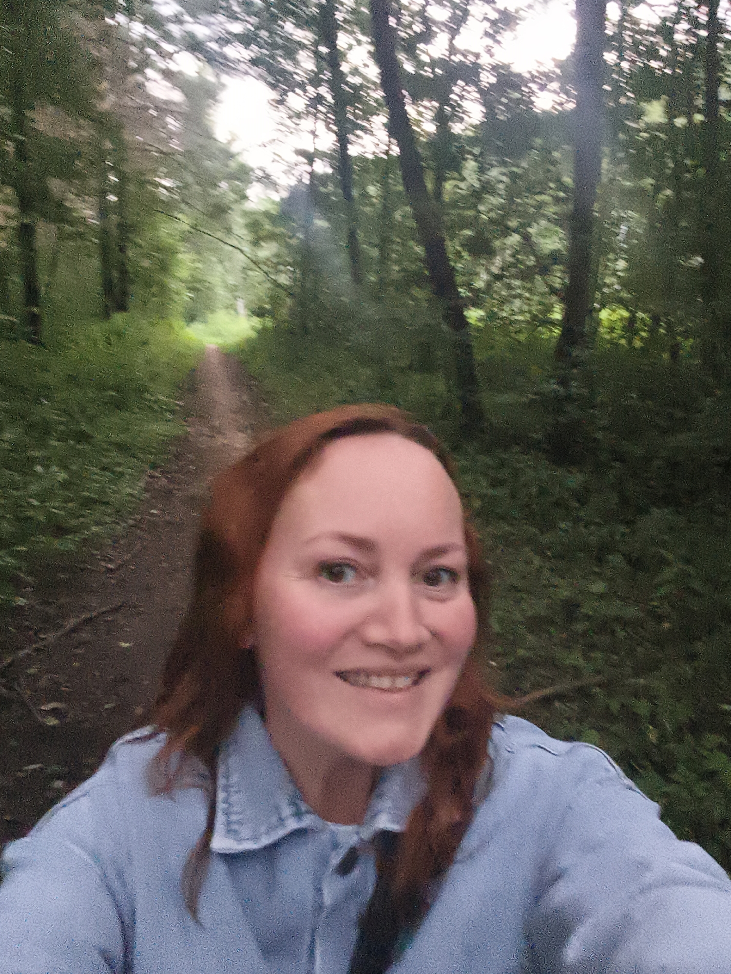 In a selfie taken from a low angle, a woman with reddish-brown hair and fair skin is smiling at the camera. She is wearing a light blue denim shirt. The background is a blurred forest path, suggesting the image was taken while walking or riding on a trail. Sunlight filters through the trees, creating dappled light. The location is identified as Chaikovsky, Russia.