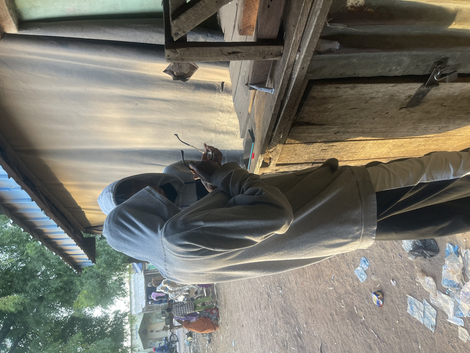 A person in a grey hoodie stands outside under a tin roof, holding glasses. Debris litters the ground, including discarded plastic. In the background, people gather, some near a building. Trees are visible beyond. An old wooden structure with a door is partially visible overhead.