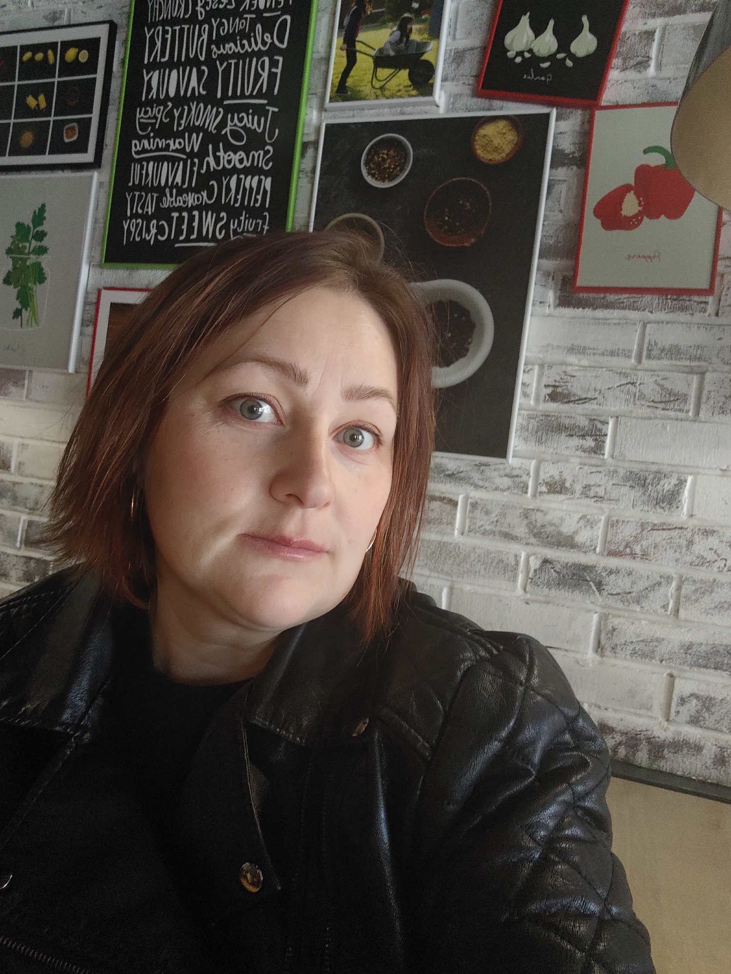 The image is a close-up, chest-up selfie of a woman taken indoors, likely in a cafe or restaurant located in Chaikovsky, Russia. The woman has fair skin, short reddish-brown hair, and light-colored eyes, looking directly at the camera with a neutral and calm expression. She is wearing a black quilted leather-look jacket over a dark top, accessorized with small, golden hoop earrings.
The background features a white-painted brick wall adorned with various framed artworks and a blackboard. The framed pictures depict food-related themes, including a grid of yellow citrus fruits, a drawing of parsley in a glass, illustrations of garlic cloves and red bell peppers, and several images of spices in bowls. A partially visible picture shows a person in a wheelbarrow. The blackboard, located behind the woman's head, displays a mirrored list of English adjectives commonly used to describe food or flavors, such as "CREAMY," "DELICIOUS," "SMOOTH," "BUTTERY," "FRUITY," "SAVORY," "SWEET," "SPICY," "SMOKEY," "TANGY," and "CRUNCHY." The decor suggests a casual and welcoming eatery environment. The lighting in the room appears soft and ambient, characteristic of an indoor setting.