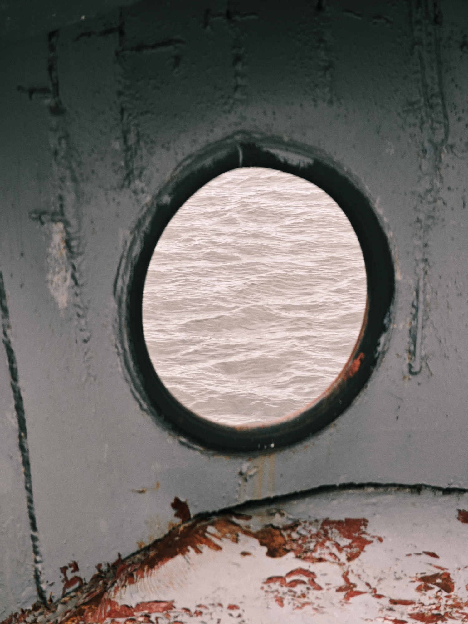 A close-up, weathered view through a porthole of a ship reveals choppy, pale pinkish-grey water. The porthole itself is circular and set within a dark, textured metal frame. The outer hull of the ship is visible in the foreground, painted a dull, dark grey with patches of rust and peeling paint, particularly along the bottom edge. The texture of the metal suggests age and exposure to the elements. The overall impression is one of a utilitarian, possibly aged vessel against a muted aquatic backdrop. The image's composition is tight, focusing on the contrast between the enclosed view through the porthole and the rough, decaying surface of the ship. No people or specific location in Zotino, Russia, are discernible from this image.