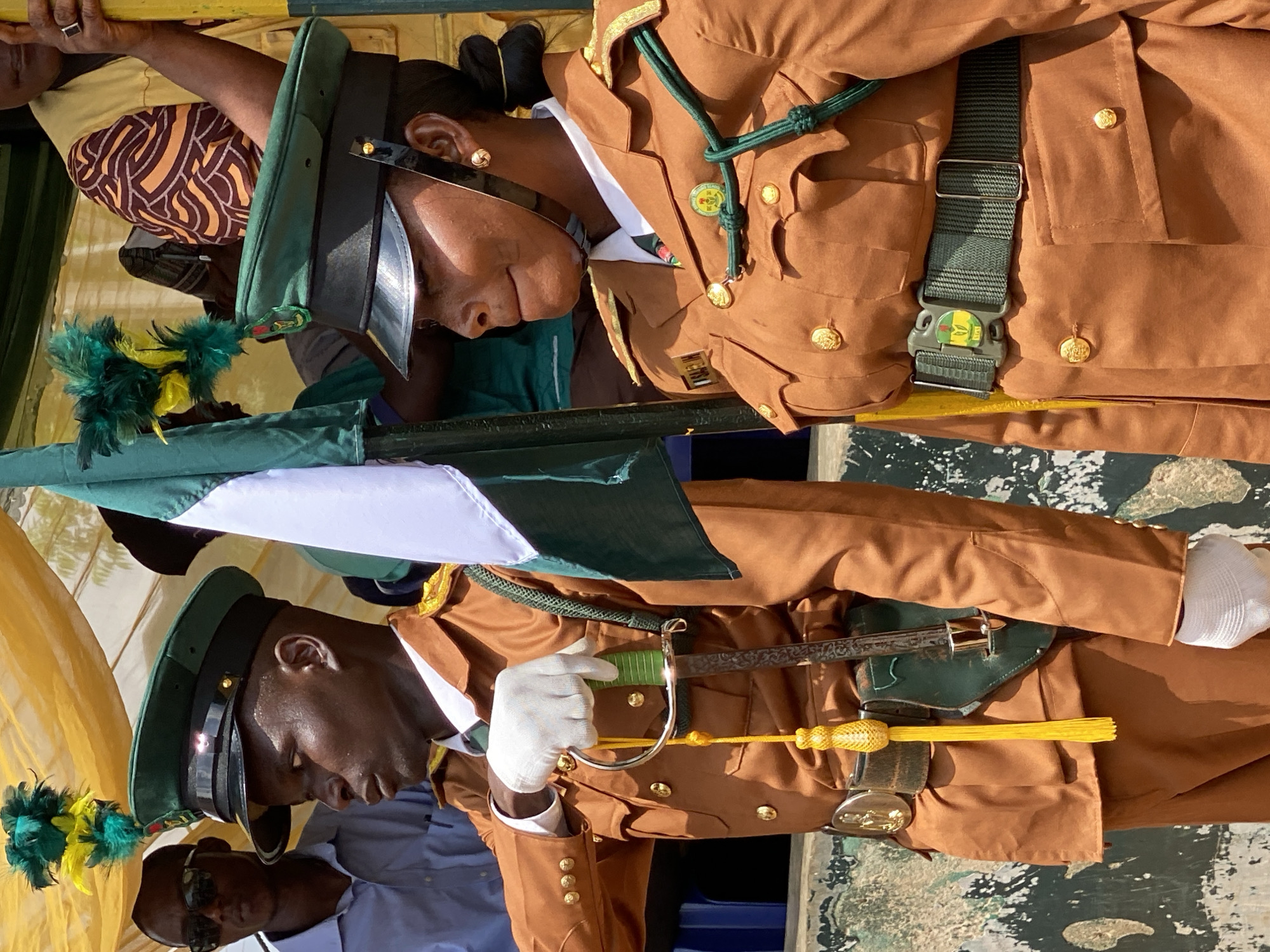 Two uniformed individuals, one male and one female, stand facing the viewer's left in front of a draped yellow fabric backdrop. The male, on the left, wears a brown uniform with a green cap adorned with feathers. He holds a sword with a green handle and a yellow tassel. The female, on the right, also wears a brown uniform with a green cap, though hers has a different brim and is adorned with a crest. A green cord is draped across her chest, and she wears a green belt with a buckle featuring a crest. Behind them, partially visible, are other individuals and a white and green flag. The overall scene suggests a formal event or ceremony, potentially in Kaduna, Nigeria, given the context of the provided crops and the nature of the uniforms.