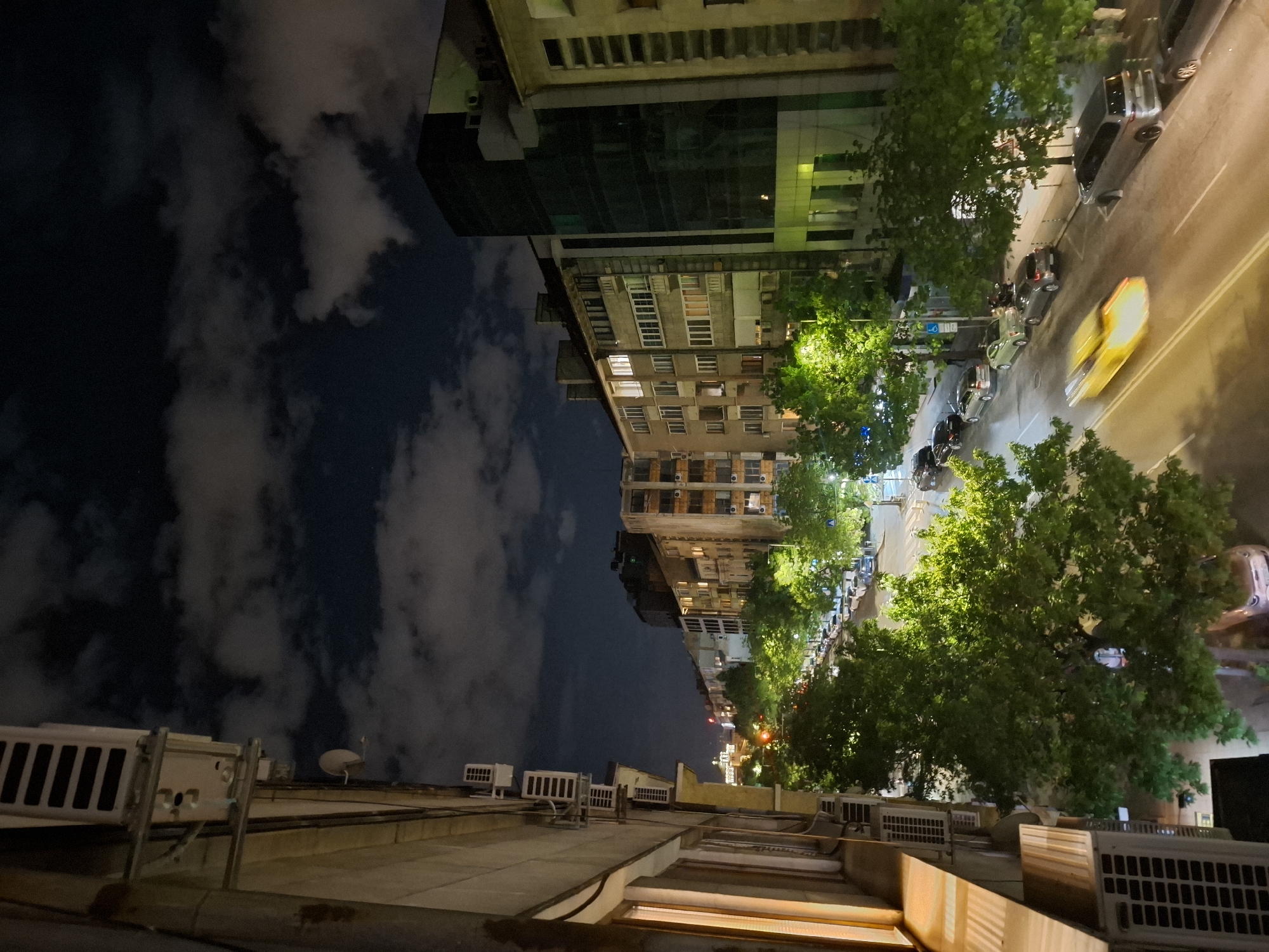 The image shows a night view in Sofia, Bulgaria. The shot is taken from a high vantage point, possibly a rooftop. The sky is cloudy. Below, a street is visible, lined with trees. A yellow car is driving along the street, causing a motion blur. Buildings flank the street on both sides. Air conditioning units are visible on the rooftops in the foreground. Cars are parked along the side of the street.