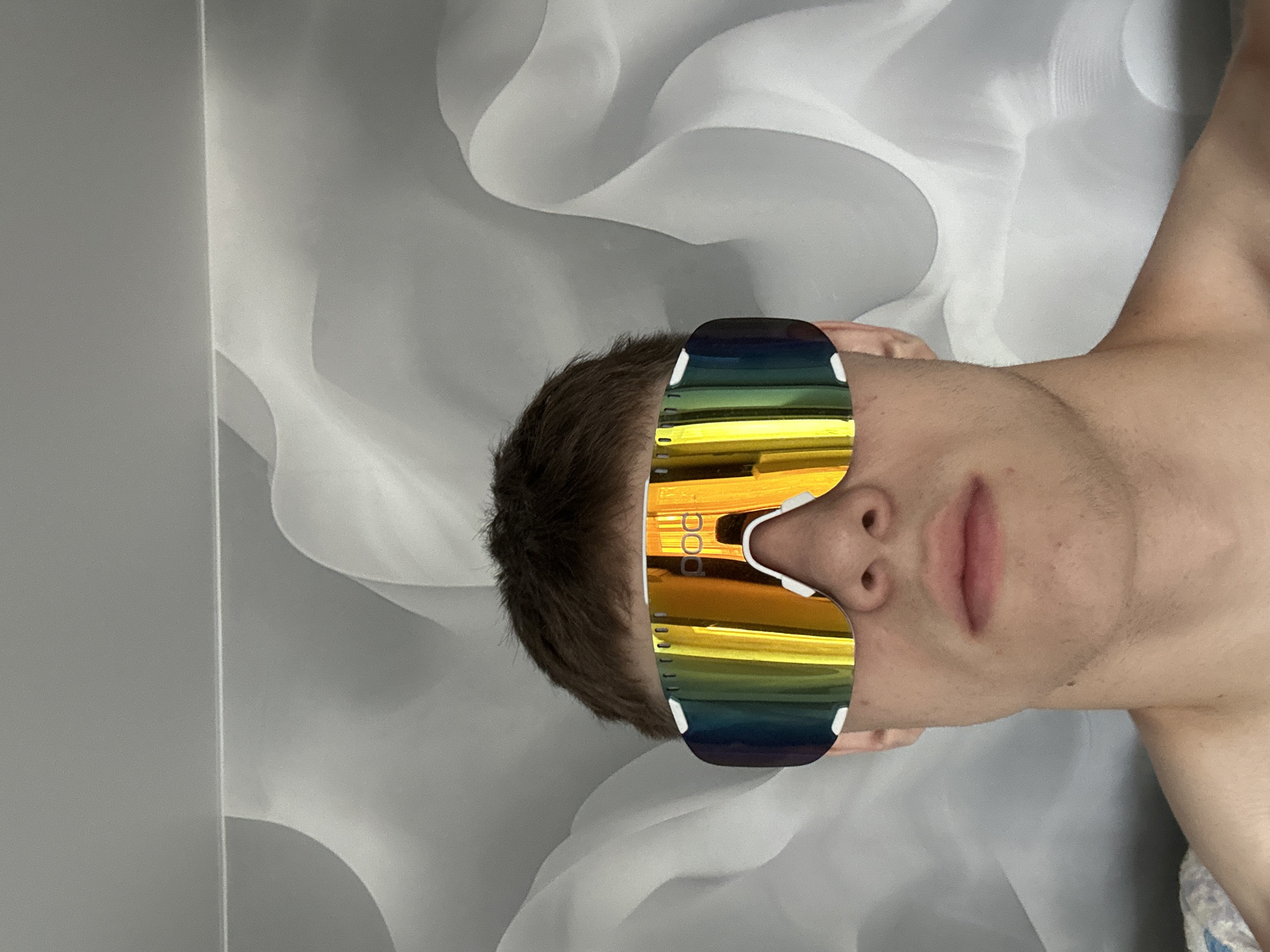 The image shows a close-up of a young person wearing large, reflective goggles with a rainbow-colored lens. The goggles have a white frame and are branded with the logo "POC." The person has short brown hair and is looking directly at the camera. They are shirtless, and only the upper chest and shoulders are visible. The background consists of a textured, draped fabric in shades of grey and white, creating an abstract, somewhat ethereal effect. The lighting is soft, illuminating the subject's face and the reflective qualities of the goggles. There is no visible indication of the specific location in Birobidzhan, Russia.