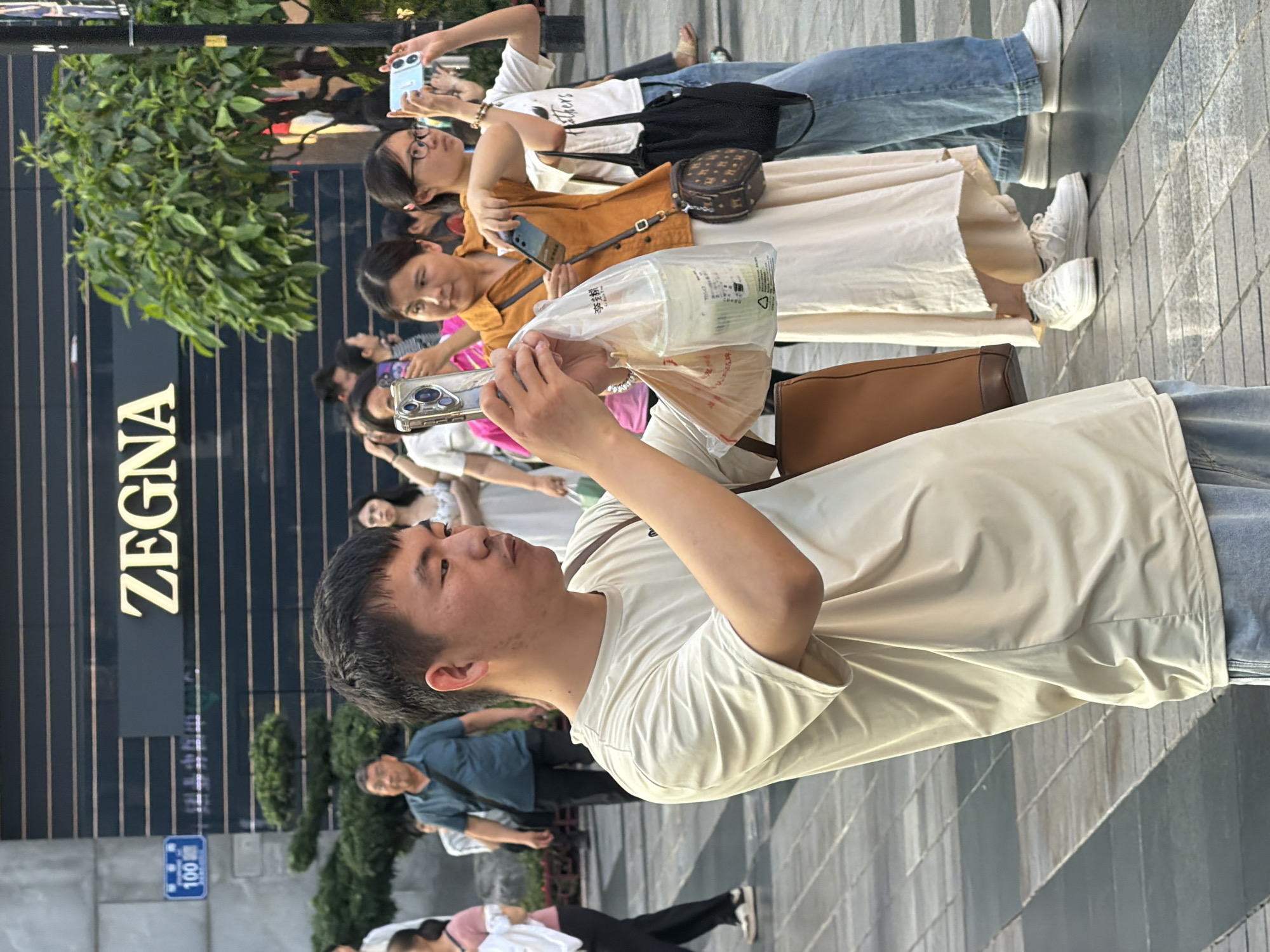 In the Yuzhong District of China, a man in a beige t-shirt and light blue jeans is standing and holding up his phone to take a picture. He is holding a plastic bag with a drink in his other hand. Behind him, a woman in a cream-colored dress is also taking a photo with her phone, and another woman in a light brown shirt and blue jeans is to her right, also holding a phone. A large green tree is visible behind them. The background features a building with the "ZEGNA" logo displayed prominently, suggesting a commercial or shopping area. Several other people are in the background, some walking and others appearing to be observing. The ground is paved with rectangular tiles.