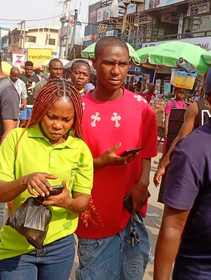 A bustling outdoor scene is captured with two people at the forefront. A woman with reddish-brown braided hair wears a lime green polo shirt and blue jeans, focused on her phone while holding a black plastic bag. Beside her, a man in a red shirt with cross designs and light blue jeans also holds a phone, a set of keys dangling from his pocket. Behind them, a diverse crowd fills the space with commercial signs like "realme" and "Infinix" visible in the background. Green umbrellas and a person carrying a container on their head add to the vibrancy of the urban scene.