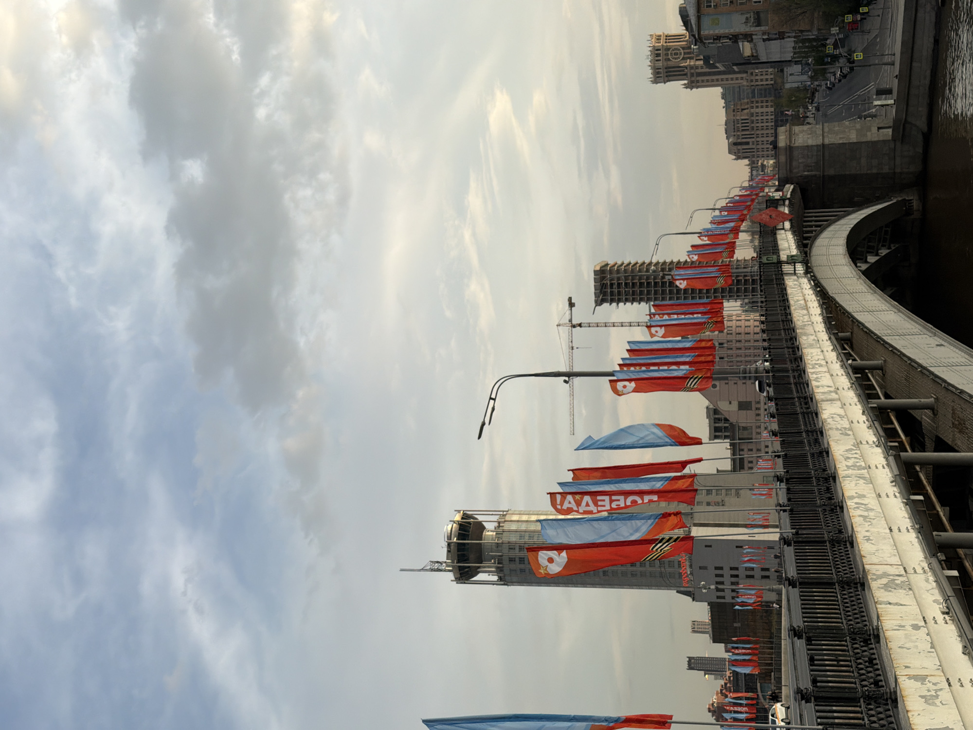 The image shows a cityscape with a bridge adorned with flags. The bridge is made of steel and features an arched structure. The flags are predominantly red and blue, bearing symbols or text in what appears to be a Cyrillic alphabet. Buildings are visible in the background, including what appears to be a skyscraper under construction with a crane. A street runs parallel to the bridge on one side. The sky is overcast with clouds. The image seems to be taken in Moscow, Russia during Victory Day celebrations due to the presence of the St. George Ribbon on the flags.