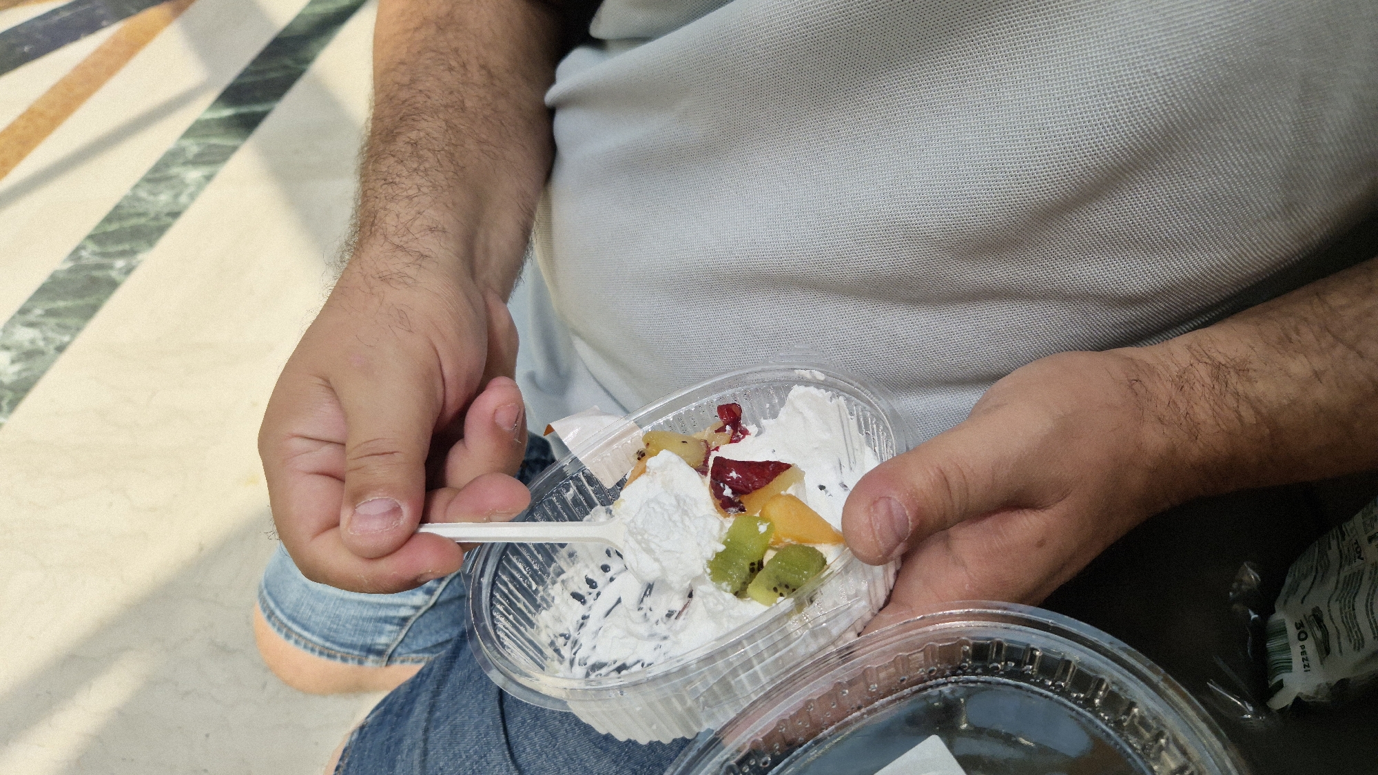 The image shows a person holding a clear plastic container of whipped cream and fruit salad. The person is wearing a light blue polo shirt and blue jeans. The container is partially filled with whipped cream and topped with pieces of fruit, including kiwi, what appears to be peach or apricot, and possibly pomegranate seeds or dried cranberries. A white plastic spoon is inserted into the whipped cream, and a piece of whipped cream and fruit has been scooped onto it.
The background shows a floor with a pattern of cream-colored and colored stripes, possibly marble tiles. The lighting suggests it is daytime. The setting appears to be indoors or in a shaded outdoor area, given the even lighting and absence of harsh shadows. The overall impression is casual, possibly a snack break or dessert. There is no visible text that is clear enough to read. The location context is Taranto, Italy.