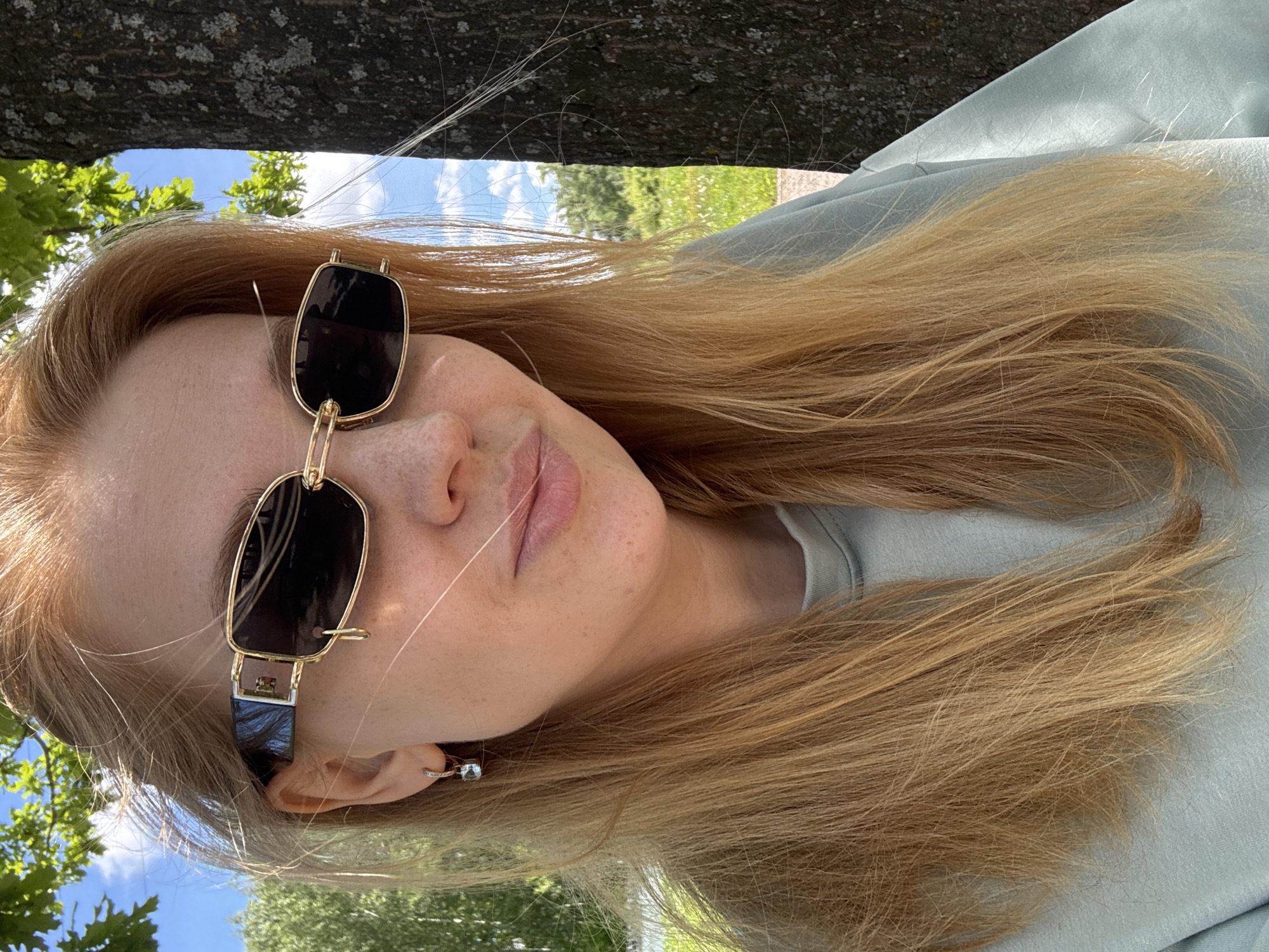 A young woman is depicted in a close-up, outdoor portrait taken in Rusanovo, Russia, under clear daylight conditions. She has long, straight, light blonde or golden-brown hair, some strands of which are illuminated by the sun, appearing almost white. Her head is tilted, and she looks towards the viewer with a relaxed expression, her lips slightly parted. Visible on her face are scattered freckles.
She is wearing large, rectangular sunglasses with dark lenses and distinctive gold-colored frames. The temple arm of the sunglasses features an intricate gold design that extends downwards towards her ear. In her visible left ear, she wears a small, delicate hoop earring. Her attire consists of a light mint green or light blue top, possibly a sweatshirt or t-shirt, which is partially visible.
The setting appears to be a natural, outdoor environment. A large, rough-textured tree trunk or branch, covered with moss or lichen, extends across the top portion of the frame, suggesting the woman is positioned beneath or near a tree. In the background, a bright blue sky with soft white clouds is visible, along with lush green foliage from other trees or bushes, indicating a park, garden, or similar open space. The bright sunlight suggests it is midday or afternoon. There is no visible text in the image.