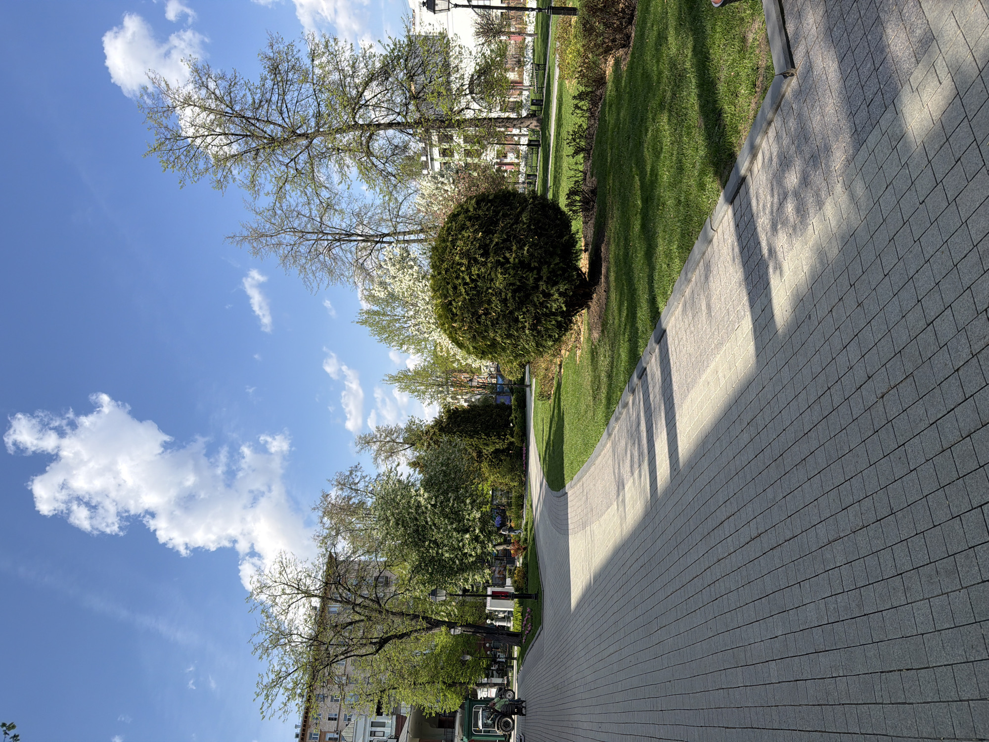 The image depicts a scene in a park in Moscow, Russia. A gray brick pathway curves through a green lawn with trees and shrubs. Some buildings are visible in the background. The sky is blue with scattered clouds.