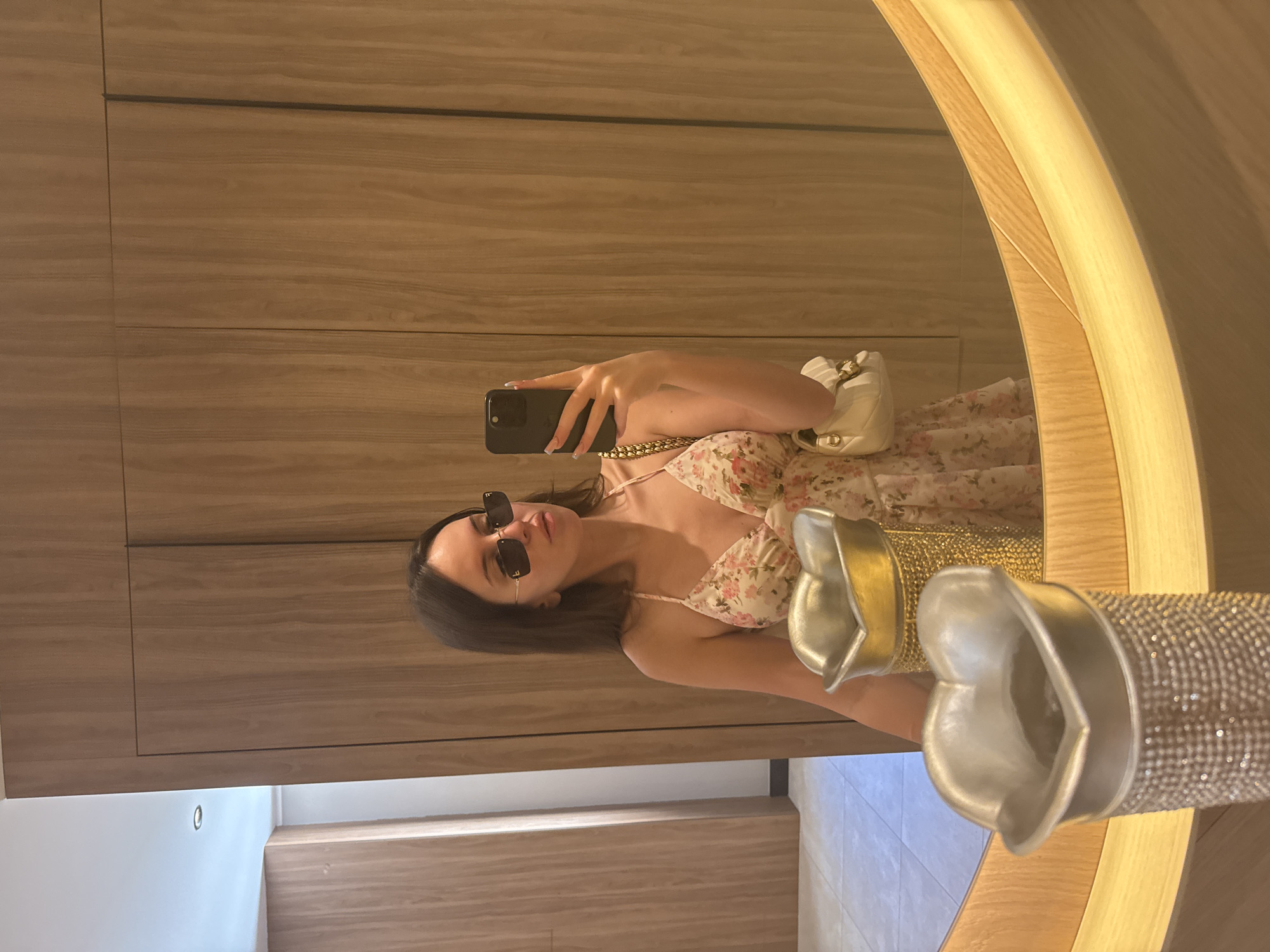 A young woman with brown hair is taking a mirror selfie in what appears to be a modern bathroom or dressing room. She is wearing a floral print dress with spaghetti straps and sunglasses. She is holding a black smartphone in her right hand to take the photo. A white purse with a gold chain strap is slung over her left shoulder. In the foreground, two decorative, heart-shaped vases are visible, one embellished with rhinestones and the other with a gold finish. The background consists of warm-toned wooden paneling and a circular mirror with integrated lighting. The overall setting suggests a private, well-appointed space.
