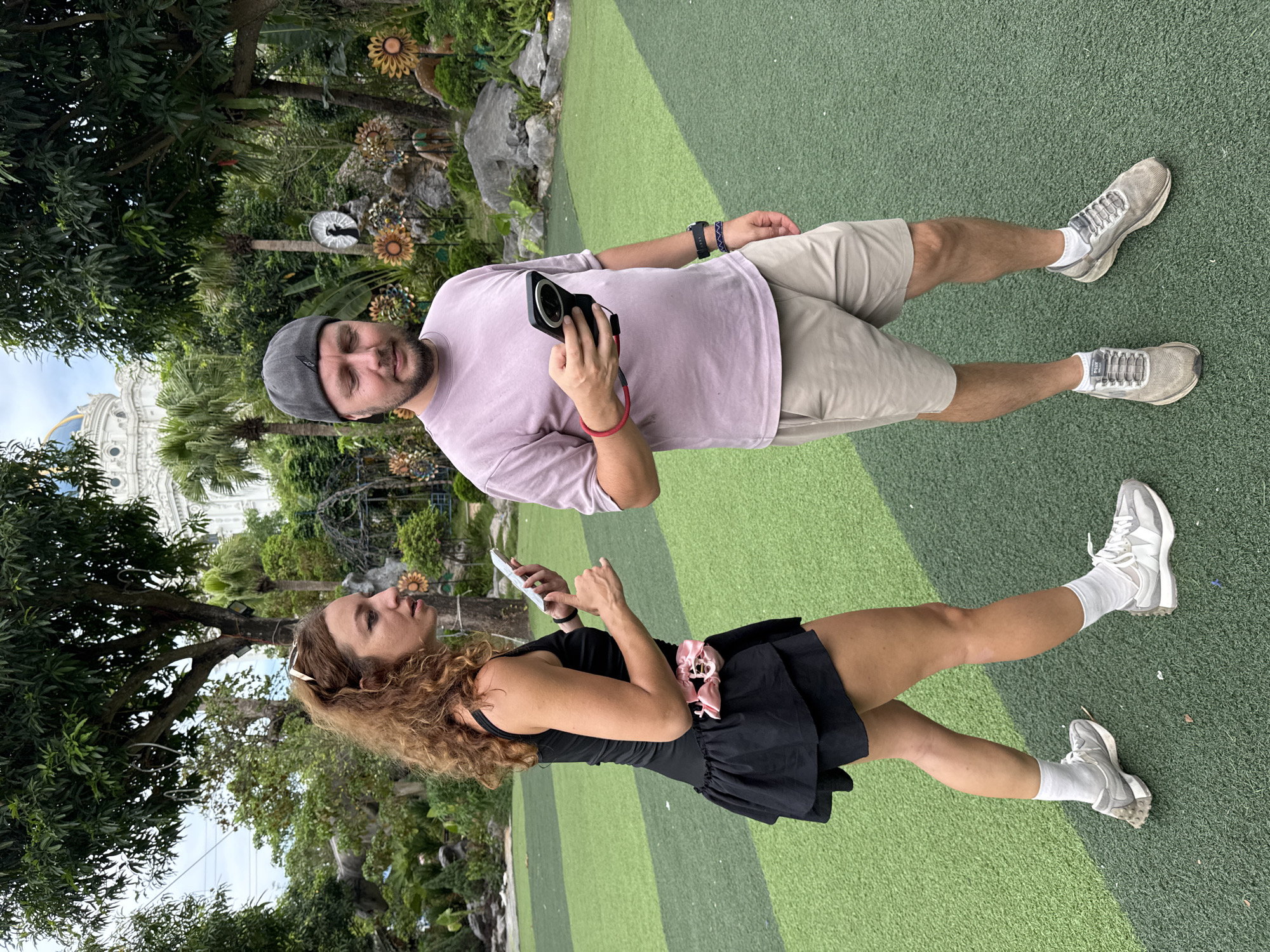 In Hoa Lư Ward, Vietnam, a woman with curly brown hair, wearing a black tank top, a black ruffled skirt, white socks, and grey sneakers, stands with sunglasses on her head. She is holding a white smartphone in her left hand and appears to be smoking. She is looking towards a man standing to her right. The man, with short brown hair and a beard, is wearing a light purple t-shirt, beige shorts, a grey baseball cap, and grey sneakers. He is looking down at a black smartphone in his right hand, which is attached to a red strap. He has a red bracelet on his left wrist and a dark blue and white patterned bracelet on his right wrist. They are standing on a green artificial turf ground with diagonal stripes. In the background, there are lush green trees, palm trees, rocks, and decorative metal flowers. A white building with a blue dome is visible in the far background.