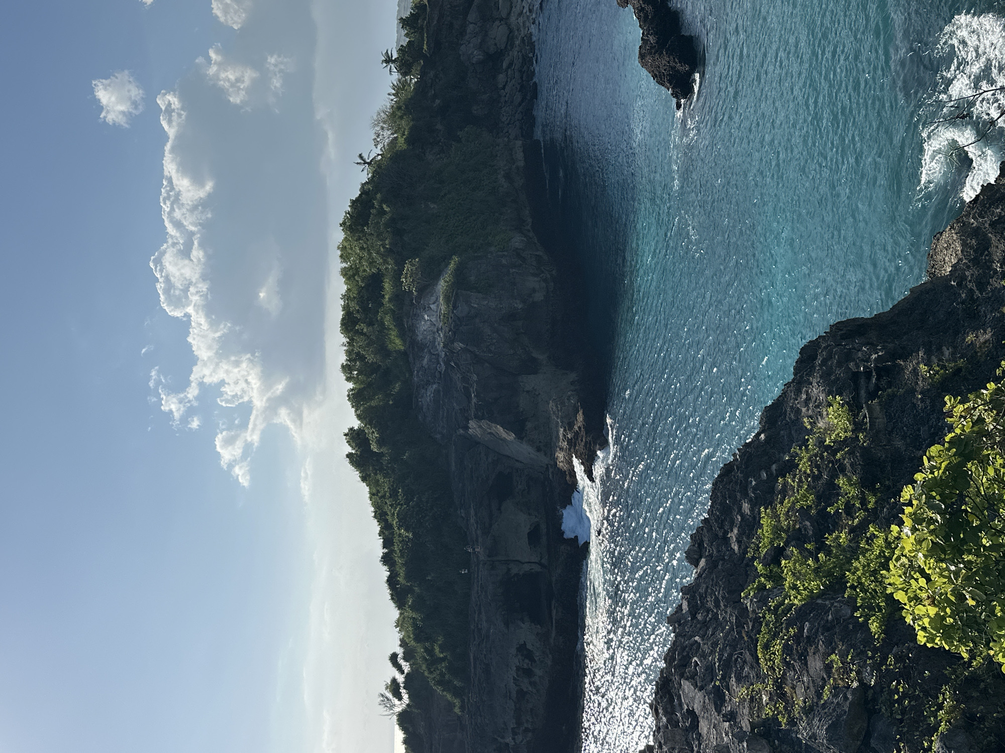 This image depicts a scenic coastal landscape in Nusa Penida, Indonesia. The scene is dominated by a rugged, verdant cliff face that plunges into the turquoise waters of the ocean. Lush green vegetation covers the top and slopes of the cliff, with some scattered palm trees visible. The sea is a vibrant blue, reflecting the sunlight, and gentle waves are seen crashing against the rocky shore, creating white foam.
In the distance, a darker rock formation juts out of the water. The sky above is a clear blue, interspersed with fluffy white clouds. The sunlight suggests it is daytime, likely morning or afternoon, given the bright illumination. There are no visible objects or text within the frame. The overall impression is one of natural beauty and tranquility, showcasing the dramatic coastline of Nusa Penida.