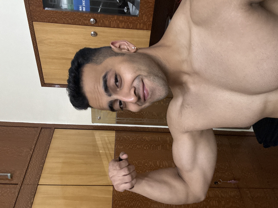 An image taken in Kolkata, India, depicts a shirtless young adult male posing indoors. The male, facing the camera directly, smiles with a slight smirk and has dark hair styled in a fade, along with visible stubble. He flexes his left bicep, with his left hand clenched into a fist. A small, gold-colored hoop earring is present in his right earlobe. The background consists of two wooden wardrobes or cabinets, featuring light brown panels bordered by darker brown frames with a textured, geometric pattern. A silver handle is visible on the upper left cabinet. On the lower left cabinet door, a small keychain with a pink charm hangs from a key in a lock. To the right, a vertical reflective surface, possibly a mirror or glass panel, partially reveals a faint reflection of what appears to be gym equipment, including text that partially reads "NATIONAL NE SYSTEM."