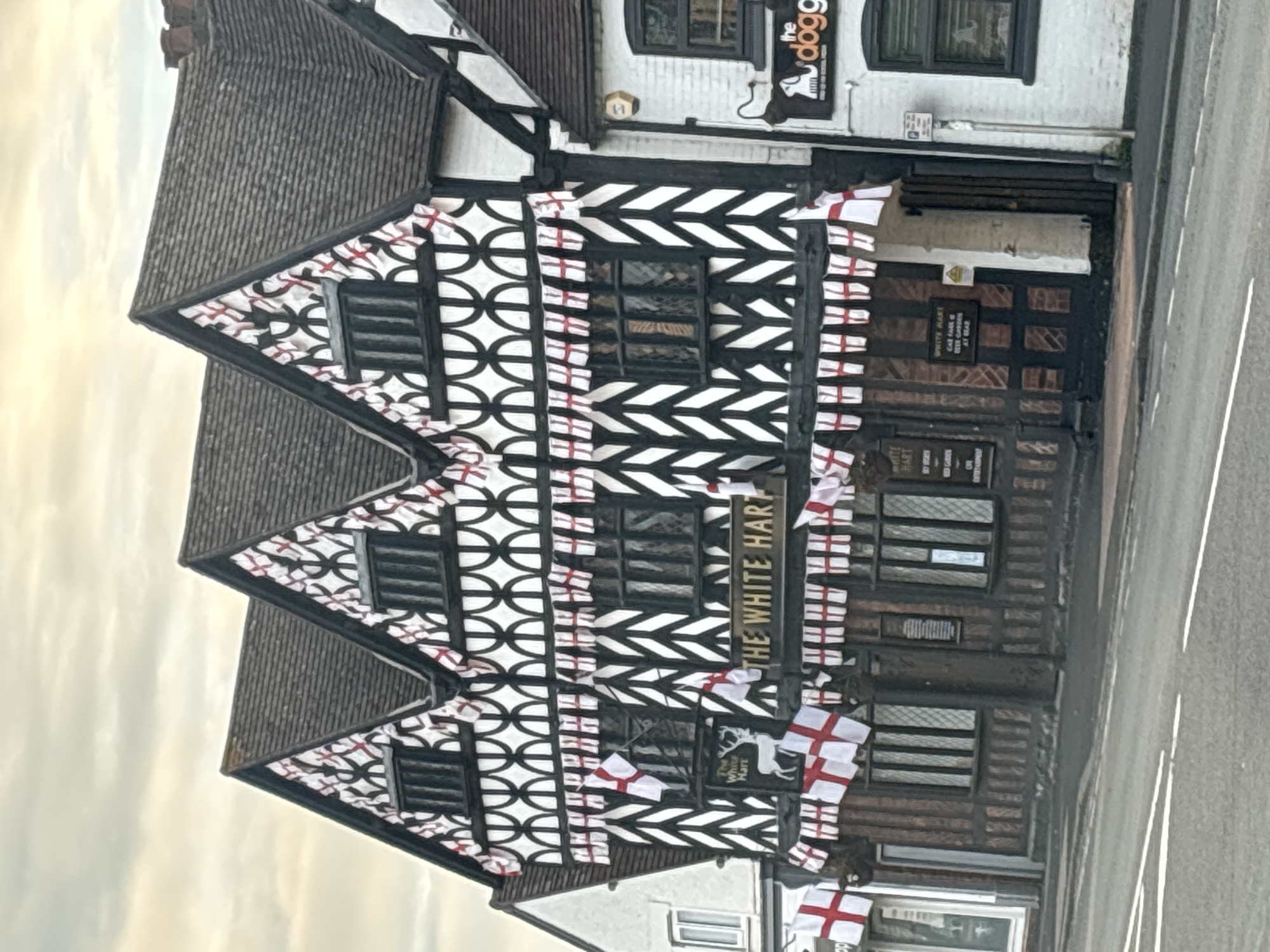 The image shows a three-story historic pub called "The White Hart" in South Staffordshire, United Kingdom. The building features a traditional black and white Tudor-style facade with decorative arches and chevron patterns. The building is adorned with numerous English flags, which are strung along the eaves and windows. The flags are red and white crosses on a white background. The pub has a prominent sign with the name "THE WHITE HART" and an image of a white stag. Adjacent to "The White Hart" is another building, painted white, with a sign that reads "the dogg." The street in front of the buildings is paved and shows white lane markings. The sky is overcast.