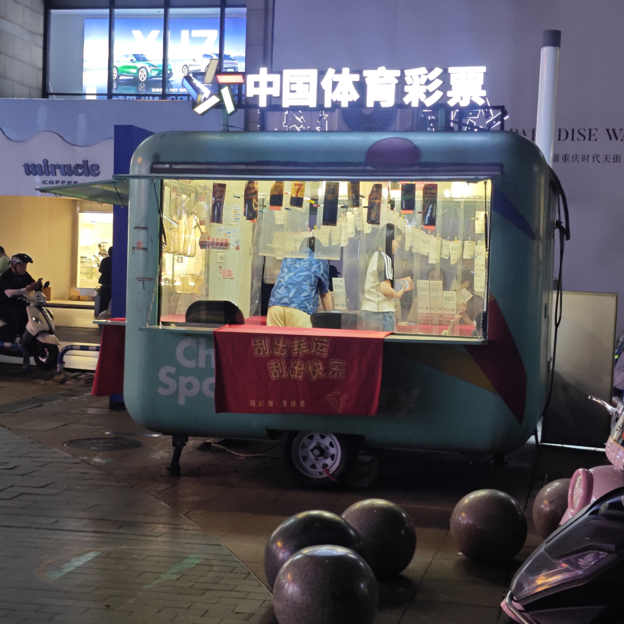 The image shows a mobile lottery stall in Yuzhong District, Chongqing, China, at night. The stall, painted a light teal color with "Ch" and "Spo" visible on the side, is brightly lit from within, displaying numerous lottery tickets hanging inside. Two people are inside the stall, one wearing a blue shirt and the other in a white and black shirt, appearing to serve customers. A red banner hangs in front of the stall with Chinese characters. Above the stall is a sign illuminated with Chinese characters. To the left of the stall is a "miracle COFFEE" sign, and above that, a billboard with cars and "X100" visible. A man on a moped is to the left of the stall. The foreground features large, spherical bollards, and parts of mopeds are visible on the right. The ground is paved with tiles. The atmosphere is urban and commercial.