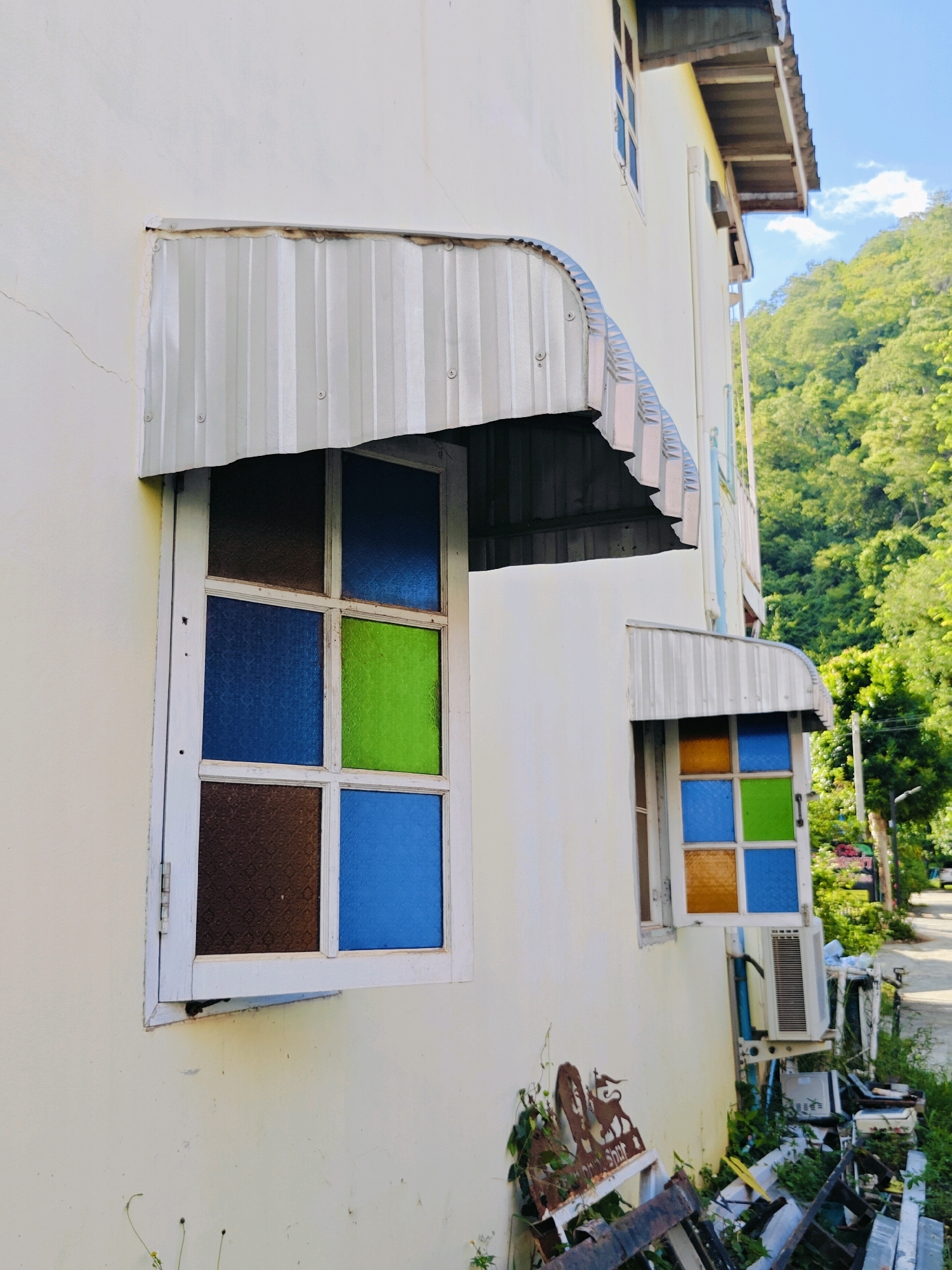 This is a photograph taken from a low angle, looking up at the side of a light-colored, two-story building in Khao Yai, Thailand. The building has a corrugated metal awning over a window on the first floor, which features stained glass panels in shades of brown, blue, and green. There is another, smaller awning and window above it on the second floor, with stained glass in orange, blue, and green. The building's wall shows some signs of wear and tear, with a crack and some discoloration near the awning. To the right of the building, lush green trees and foliage dominate the background, with a hint of a road and a street light visible in the distance. An air conditioning unit and some discarded metal objects are situated at the base of the building. The sky is bright blue with scattered white clouds.
